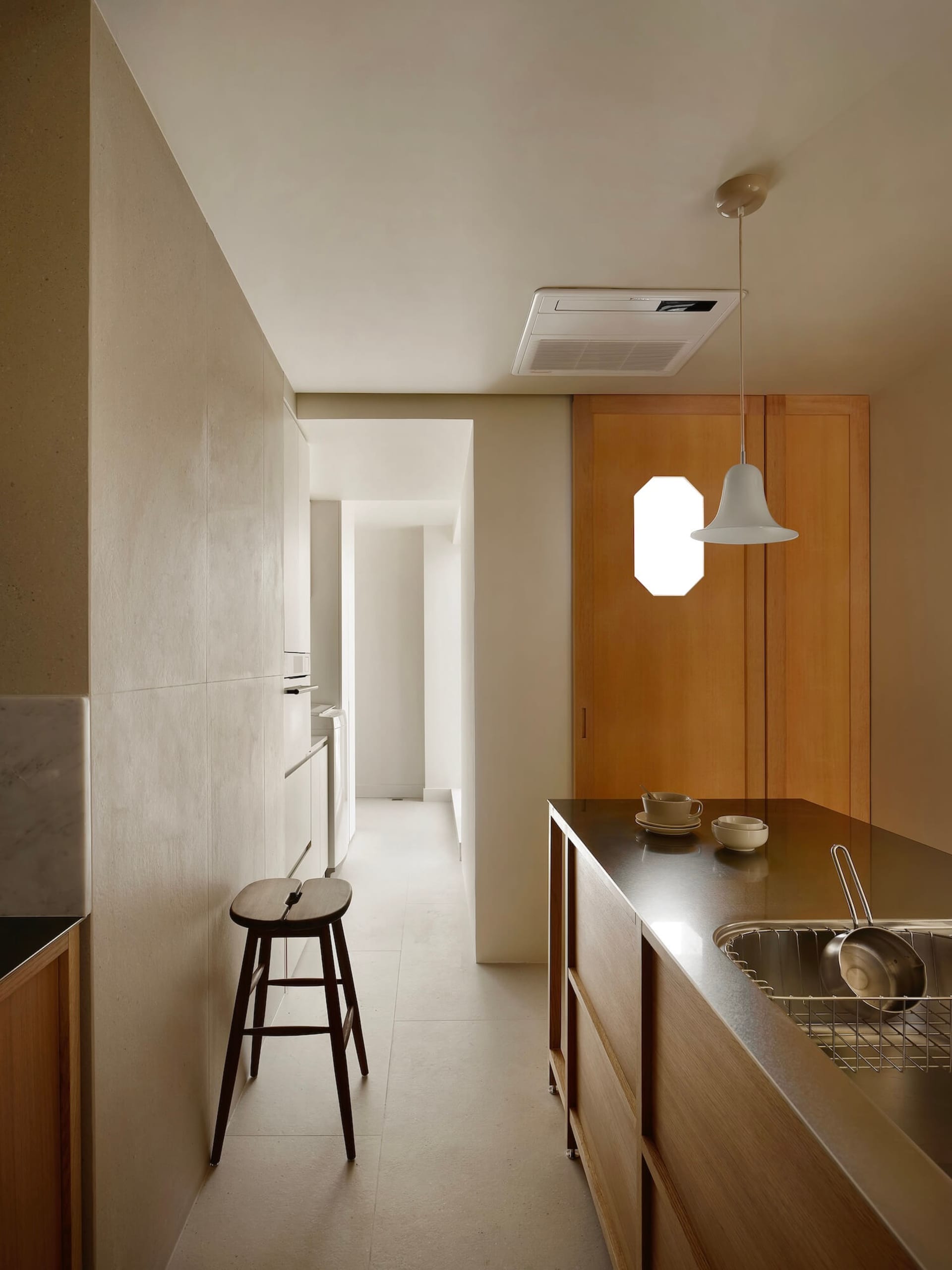 Modern minimalist narrow kitchen with light oak wood cabinetry, dark countertop, stainless steel sink, wooden stool, and white pendant light hanging from the ceiling.