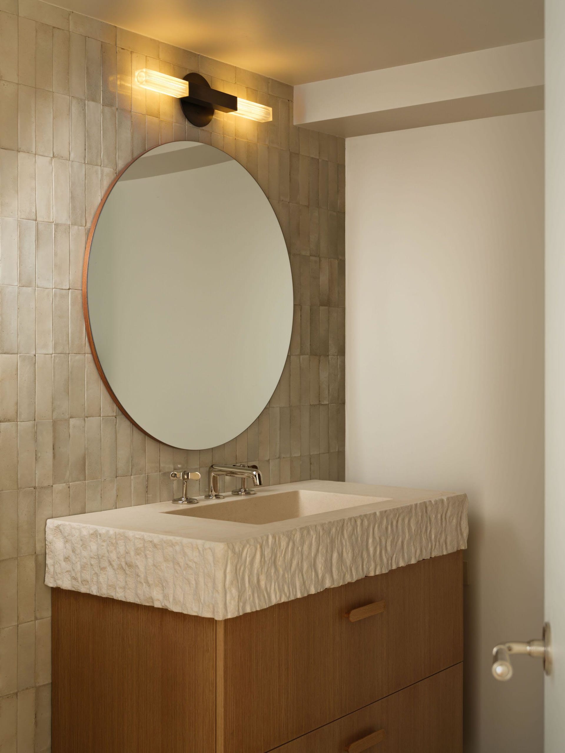 Modern bathroom vanity with a thick, textured travertine countertop, wooden drawers, a large round mirror, and a contemporary linear wall sconce emitting warm light against a beige tiled wall.