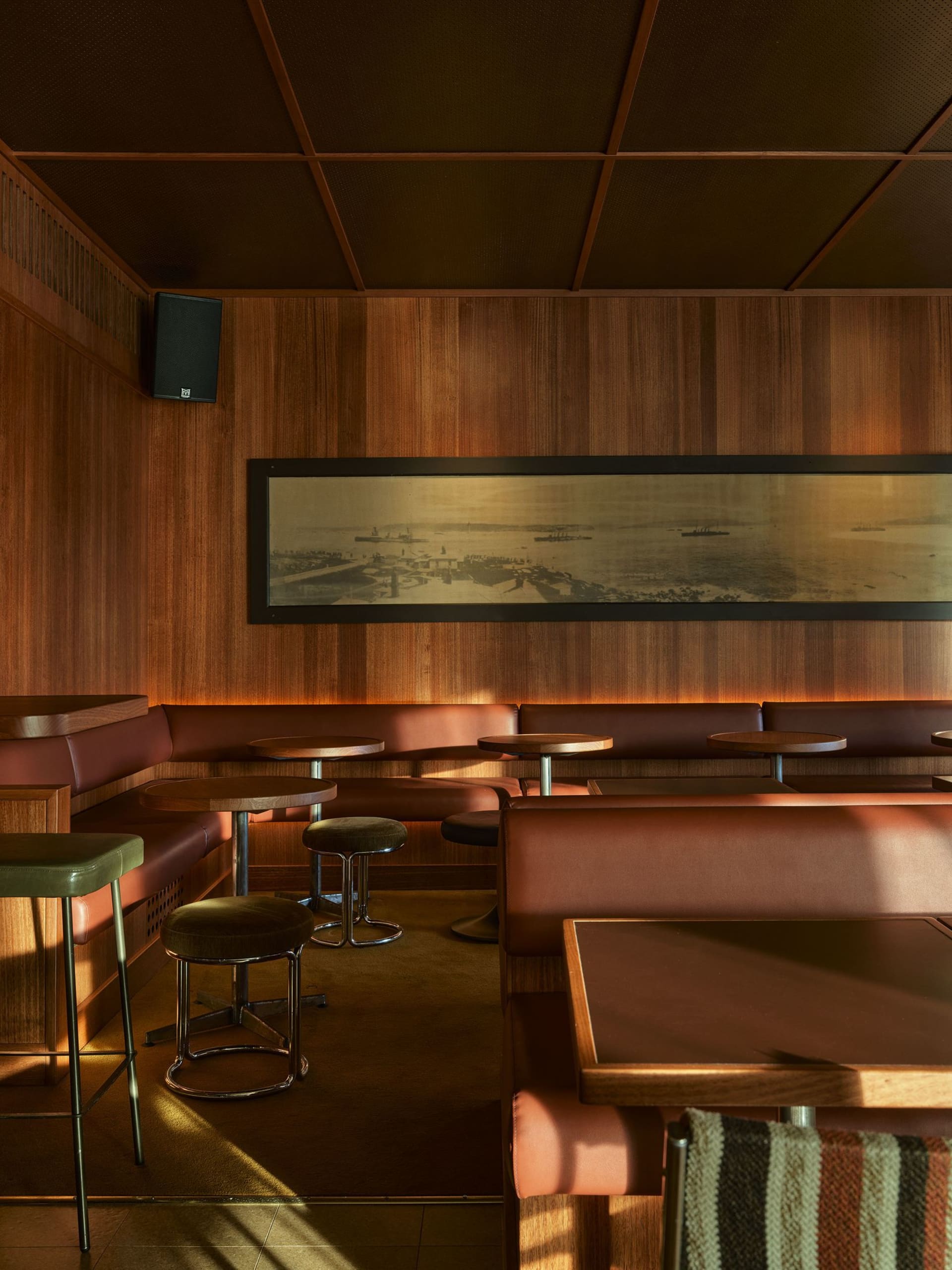 Interior view of a mid-century style bar lounge featuring walnut wood paneling, oxblood leather banquettes, round wooden tables, and a large framed harbor photograph.