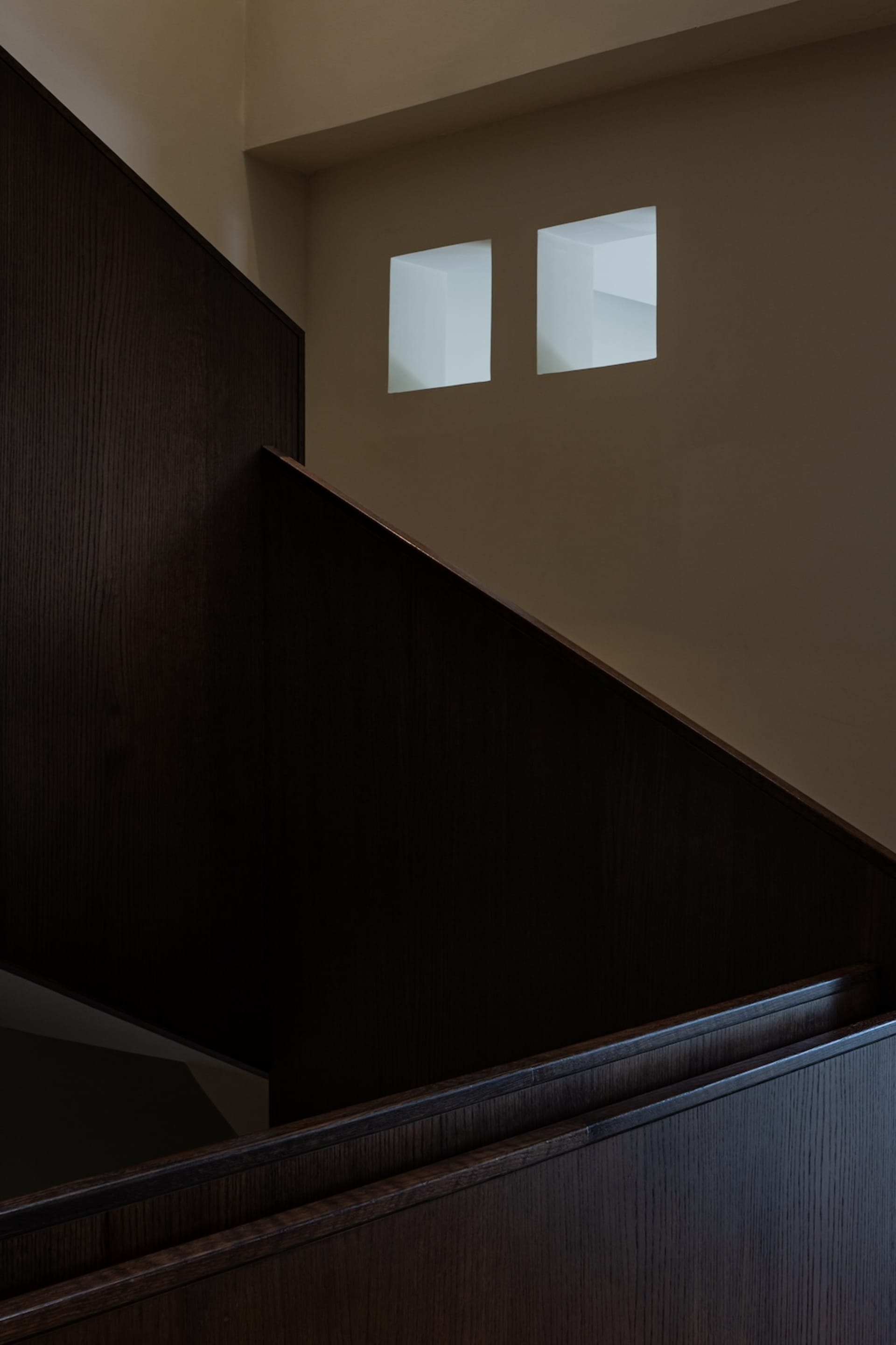 Close-up view of a dark oak wooden staircase with thick handrail and paneling, featuring two square recessed light niches in the upper beige wall.