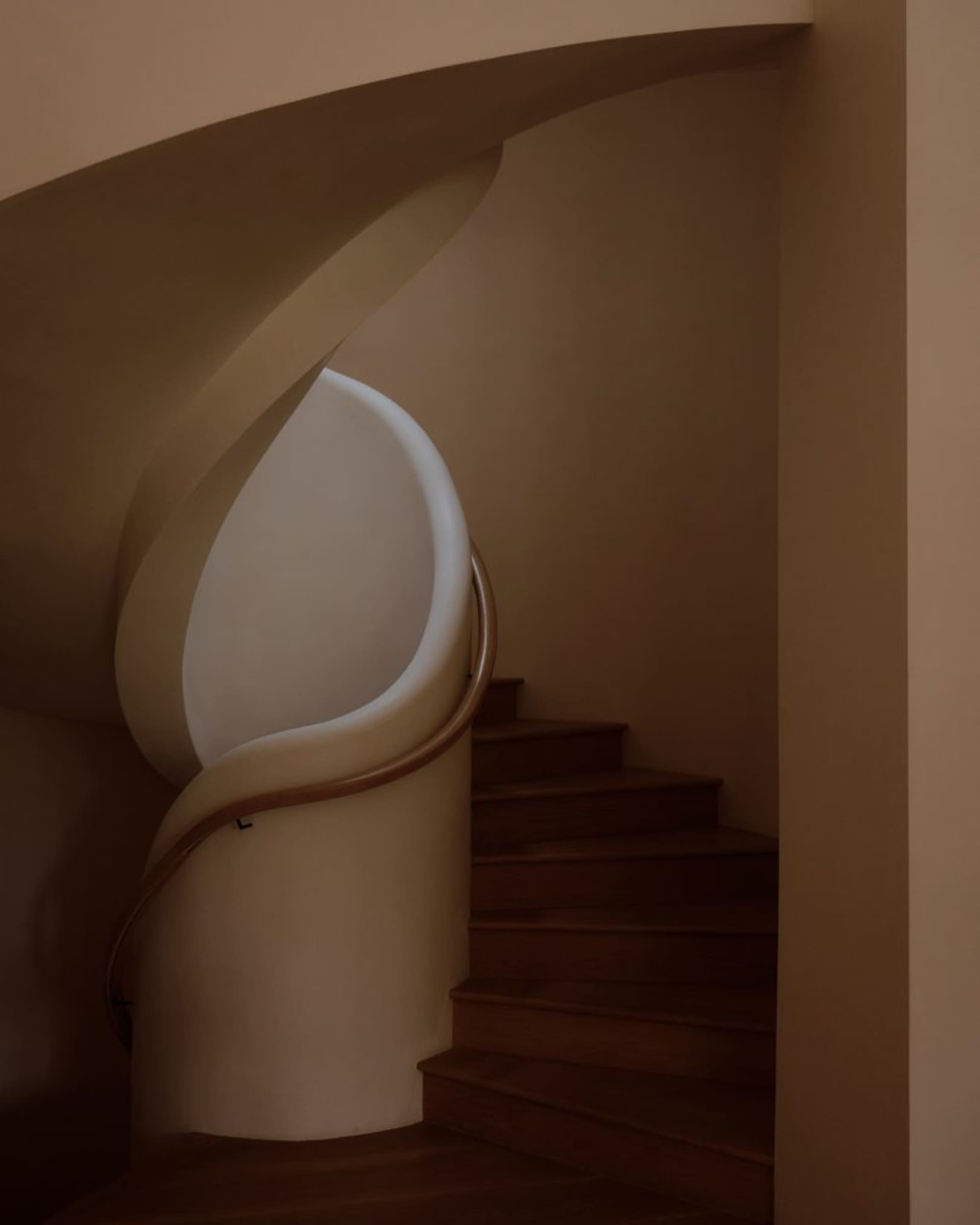 View of a modern curved staircase featuring a smooth white plaster balustrade, warm wooden handrail, and dark walnut treads ascending into soft beige walls.