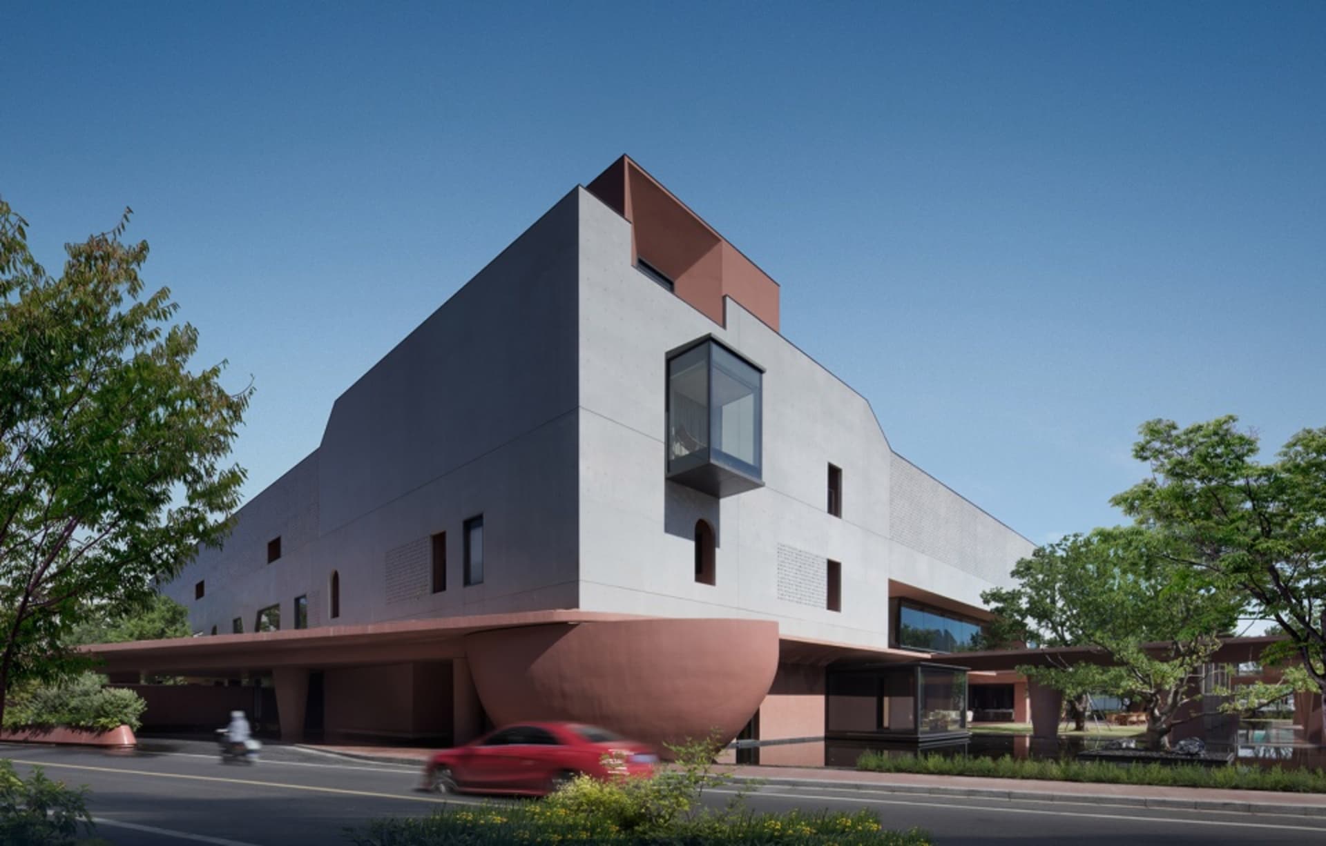 Exterior view of a modern Brutalist-style cultural center building with grey concrete upper volumes and a curved terracotta-colored base, surrounded by green trees under a clear blue sky.