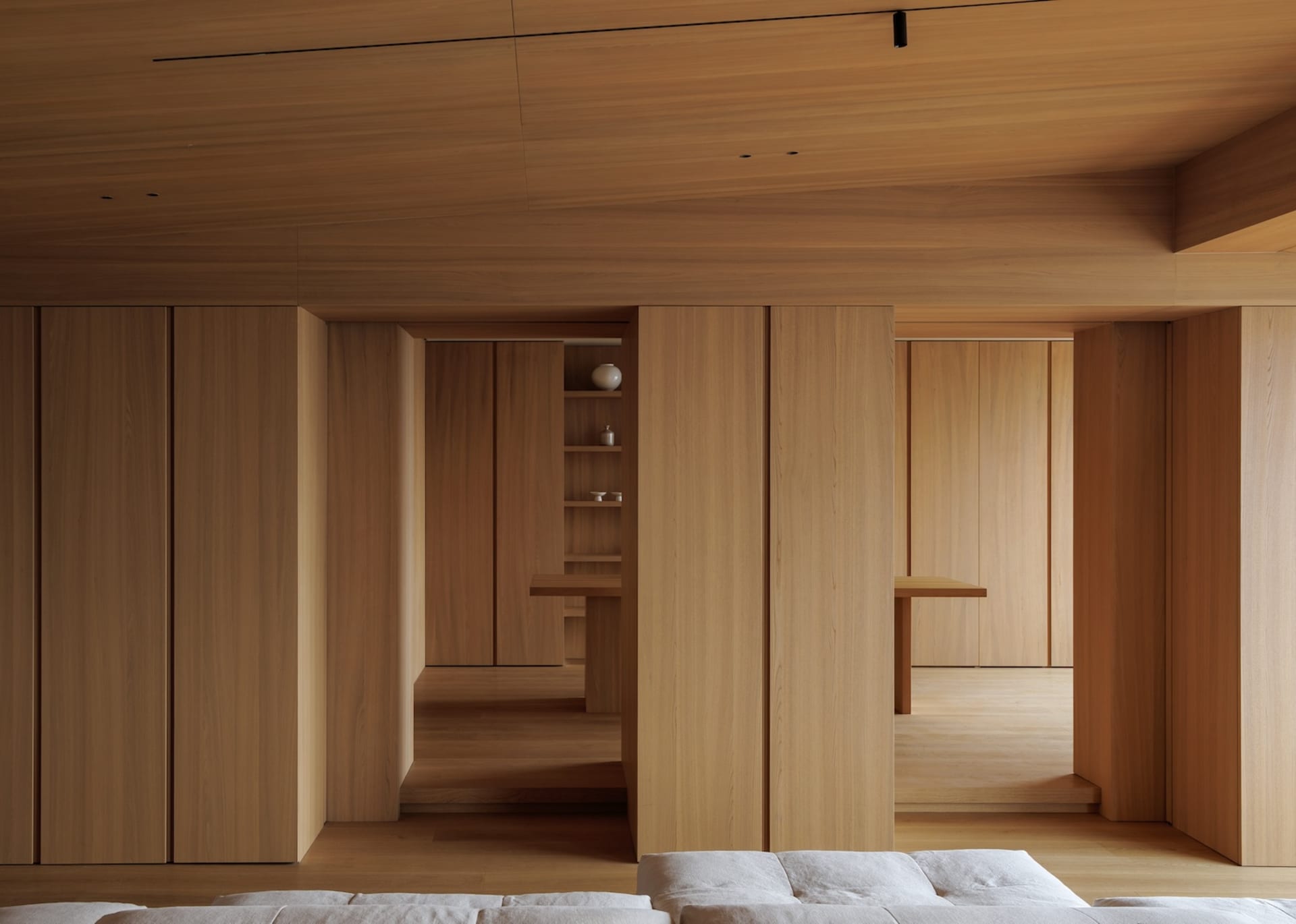 Modern minimalist living room interior featuring extensive light oak wood cabinetry, built-in shelving, and a white fabric sofa in the foreground.