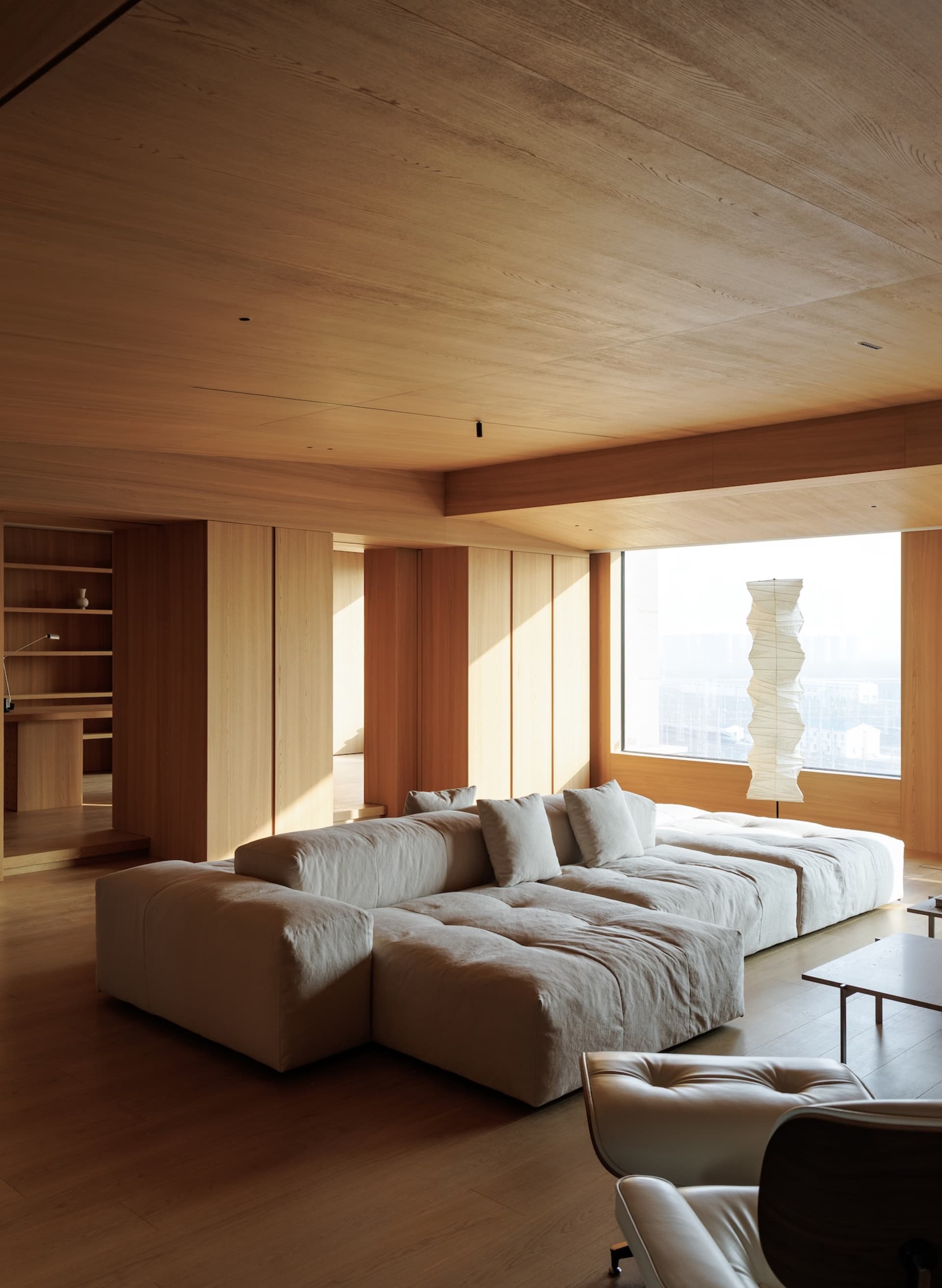 Modern minimalist living room featuring a large low-profile modular sofa in off-white fabric, extensive light oak wood paneling on walls and ceiling, a sculptural paper floor lamp, and leather lounge chairs in the foreground.