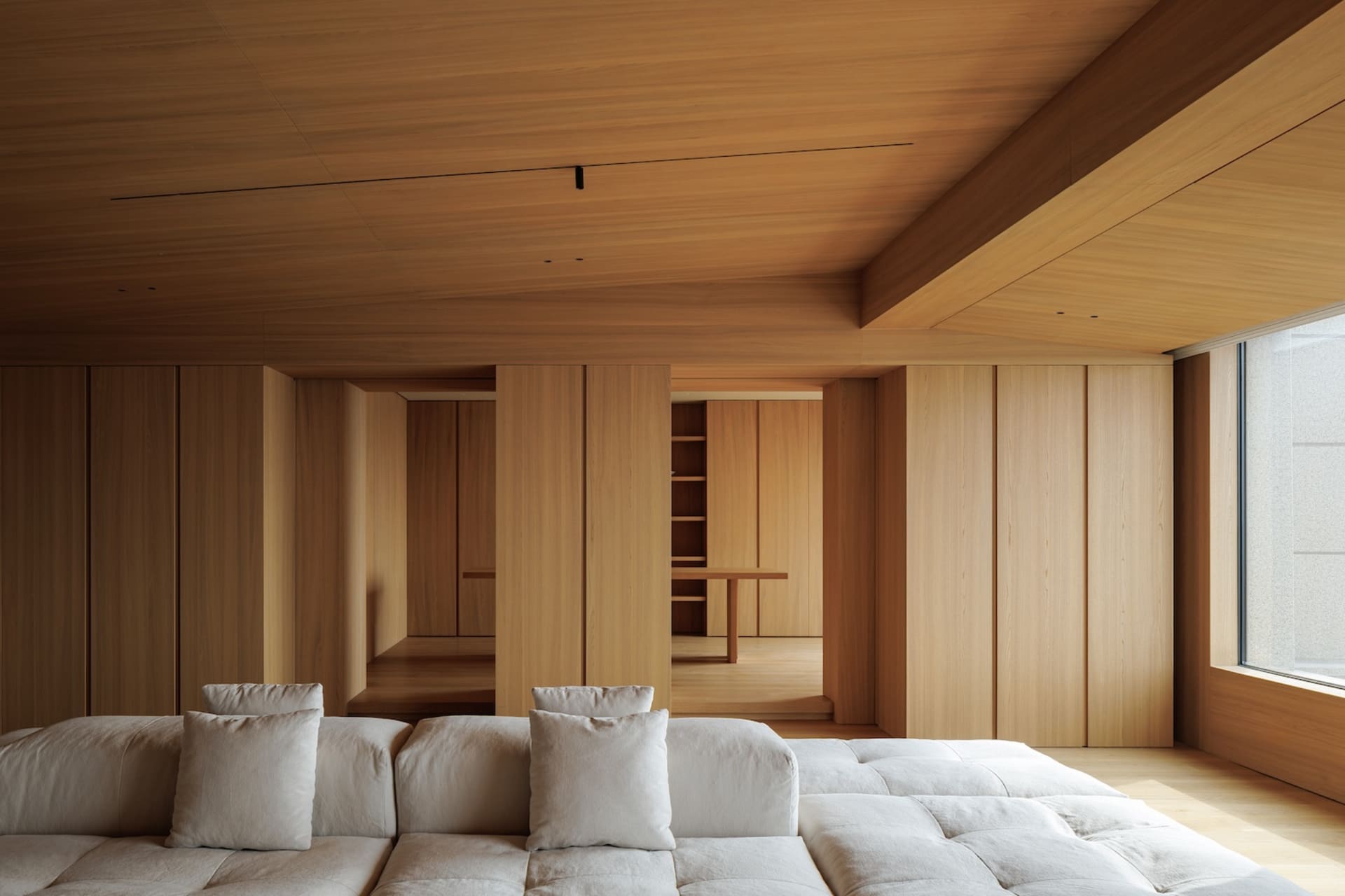 Modern minimalist living room featuring a large modular white sofa, extensive oak built-in cabinetry, wooden ceiling, and a view into a dining area.