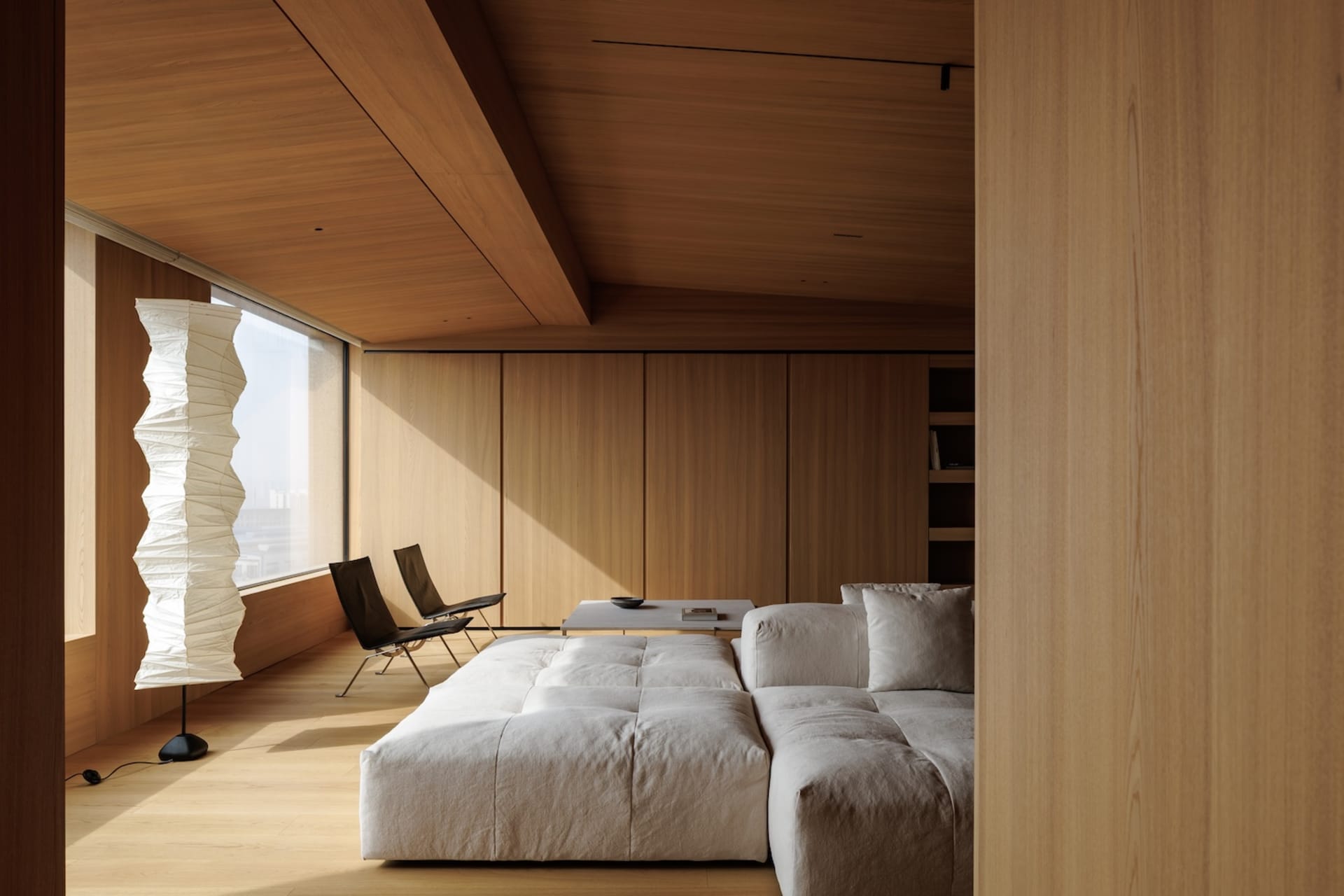 Modern minimalist living room featuring a large low-profile light gray sectional sofa, wooden ceiling and wall paneling, a tall white Akari-style paper floor lamp, and two black leather lounge chairs by the window.