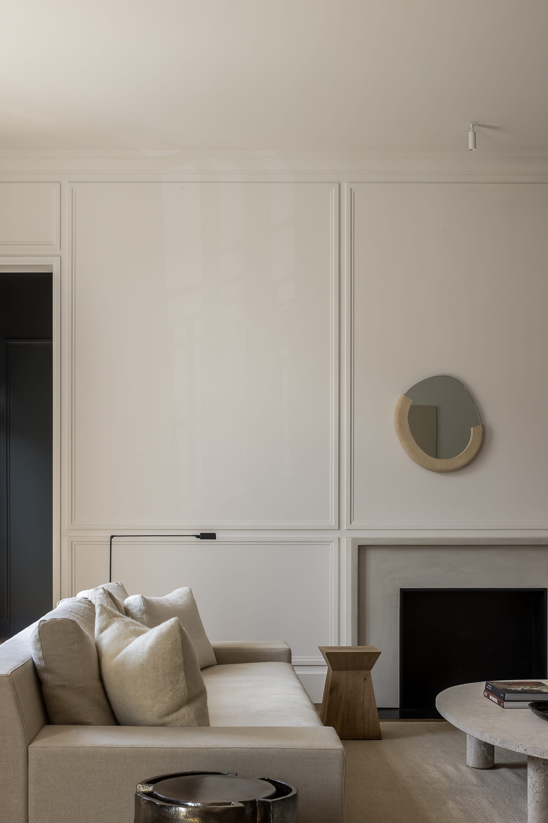 Modern minimalist living room featuring a beige linen sofa with plush cushions, a black fireplace framed in limestone, an irregular organic-shaped mirror with a textured beige frame, and a round concrete coffee table on a light wood floor.