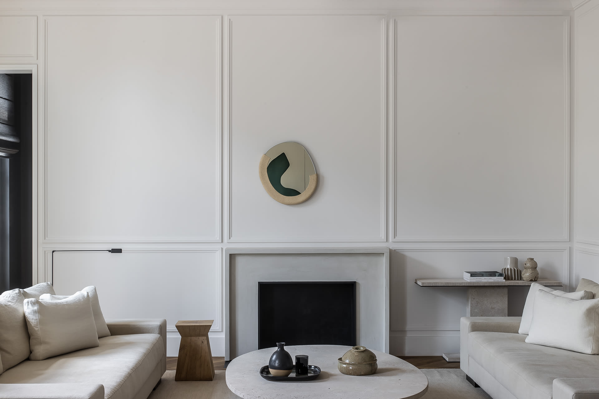 Minimalist living room with two cream sofas facing a round concrete coffee table, a black fireplace with limestone surround, and an irregular organic mirror on a white paneled wall.