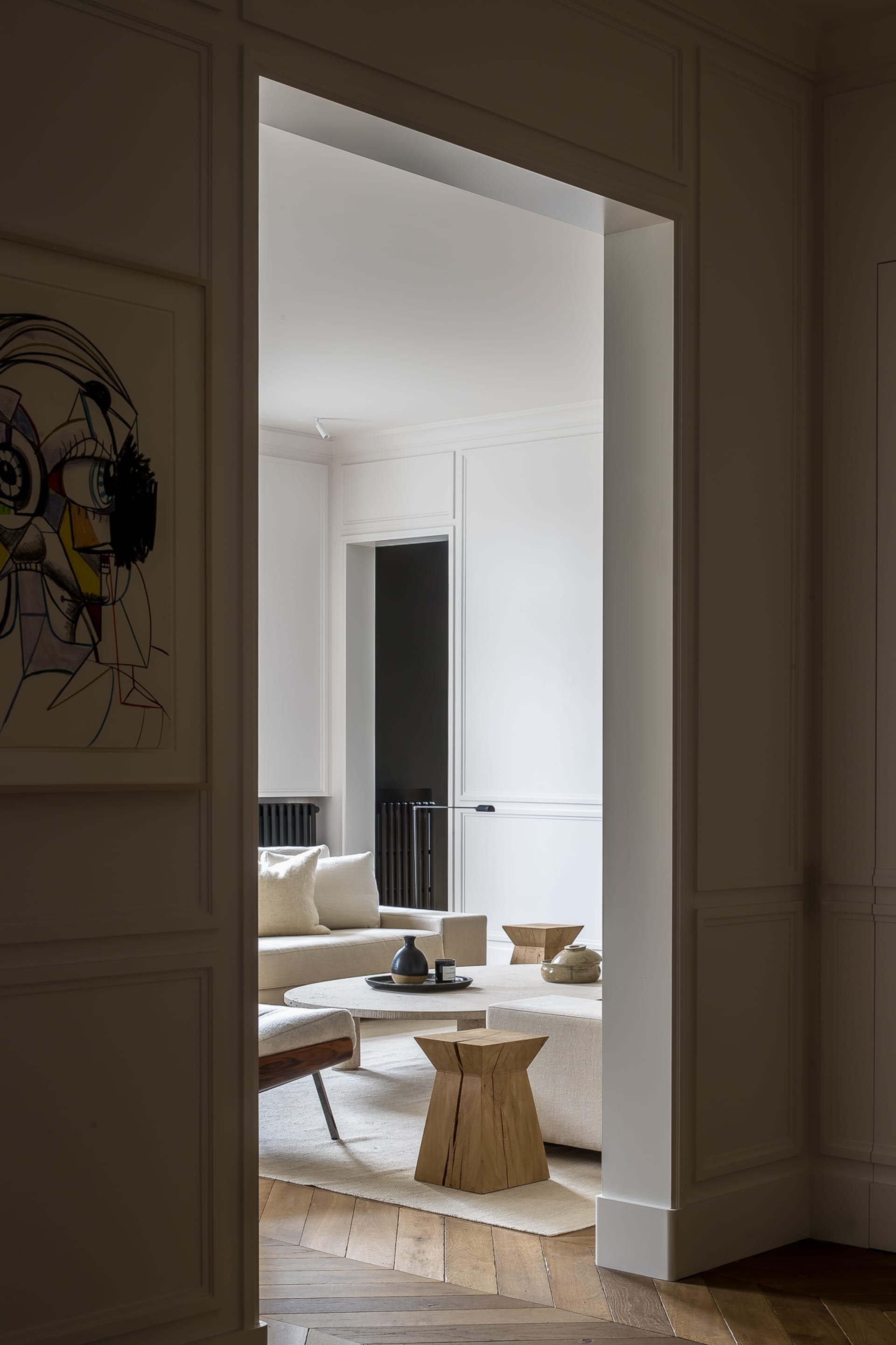View through a doorway into a minimalist living room featuring a cream linen sofa, sculptural oak side tables, a round travertine coffee table, and a large abstract portrait painting.