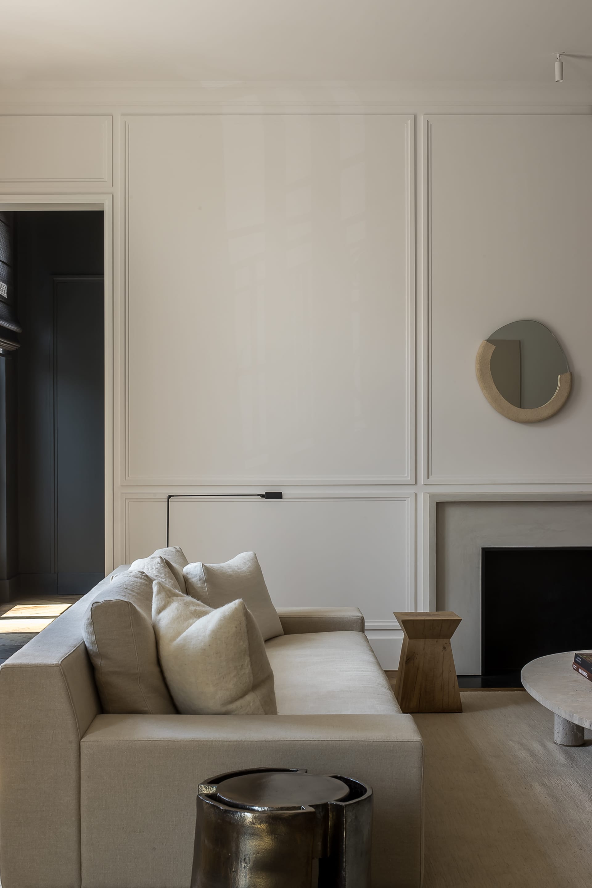 Modern neutral living room featuring a beige linen sofa with plush pillows, a sculptural oak side table, a metallic coffee table, and an irregular organic mirror against white panelled walls.