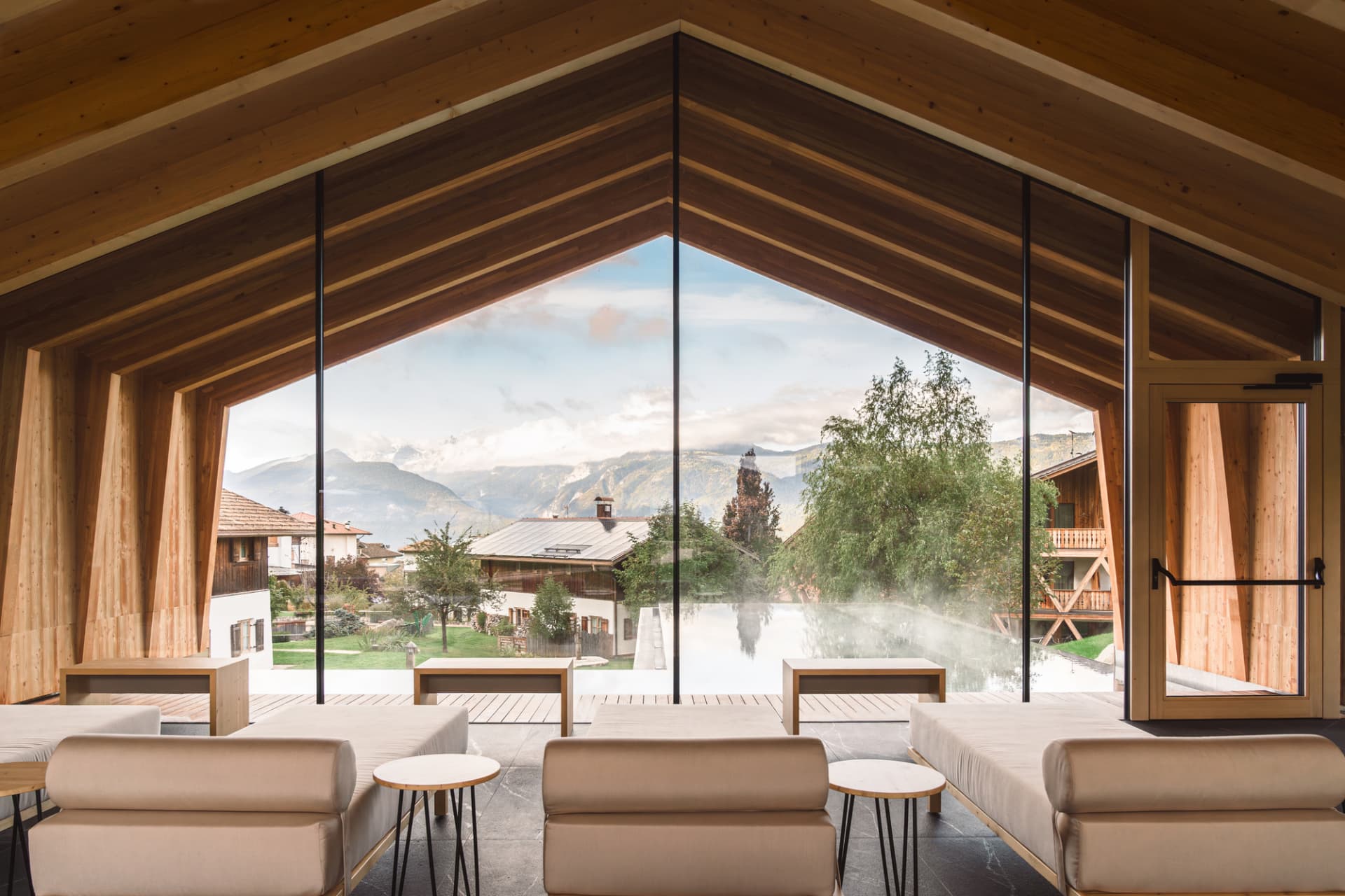 Modern wooden lounge with beige sofas facing large triangular windows overlooking a steaming pool and alpine mountains