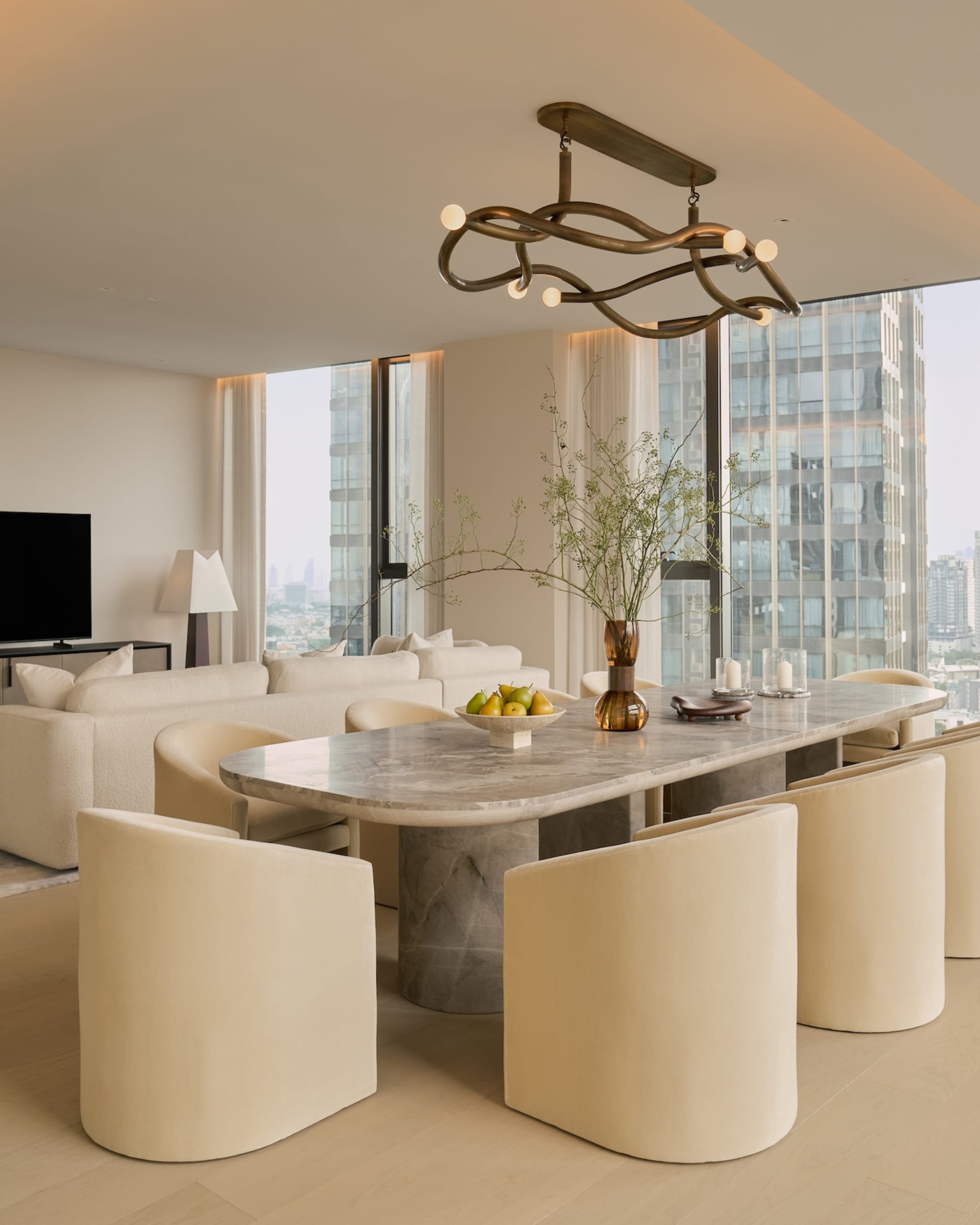 Modern luxury dining area featuring a large Calacatta marble table, six cream bouclé curved chairs, and an artistic bronze chandelier with floor-to-ceiling city views in the background.