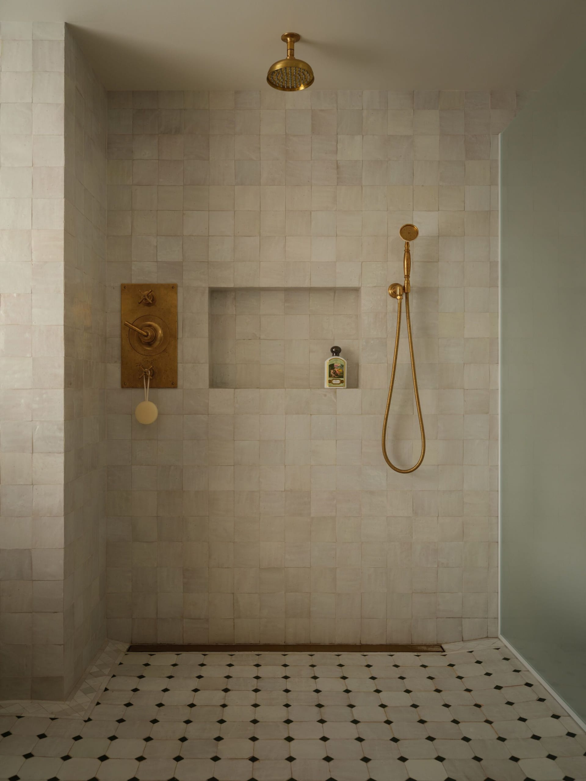 Modern luxury shower corner with light marble tiled walls, recessed niche, unlacquered brass shower head and valve, and geometric black and white marble floor tiles.