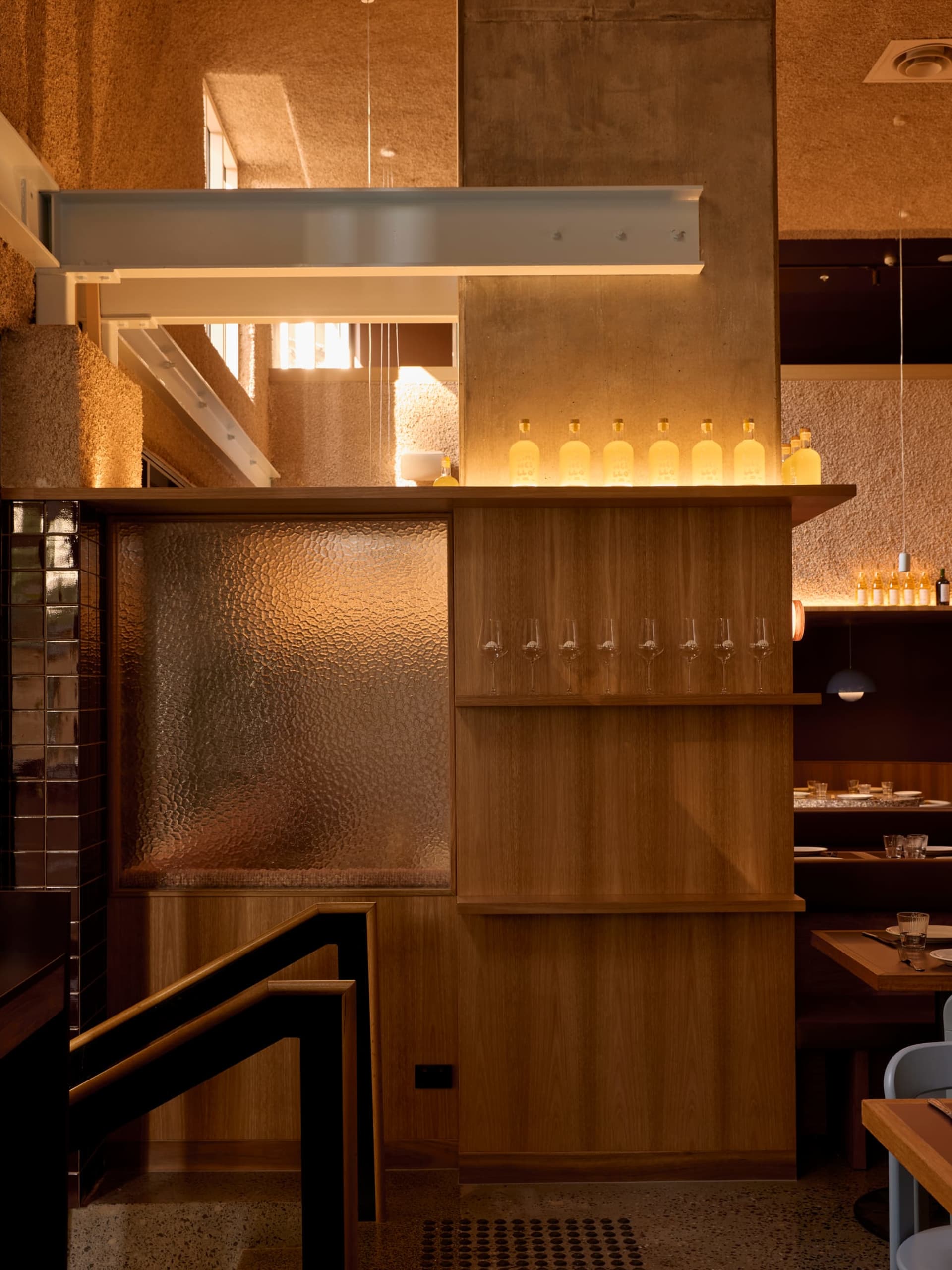 Modern restaurant bar area featuring textured plaster walls, warm wood cabinetry, backlit bottle display, and a hammered metal panel under dramatic warm lighting.