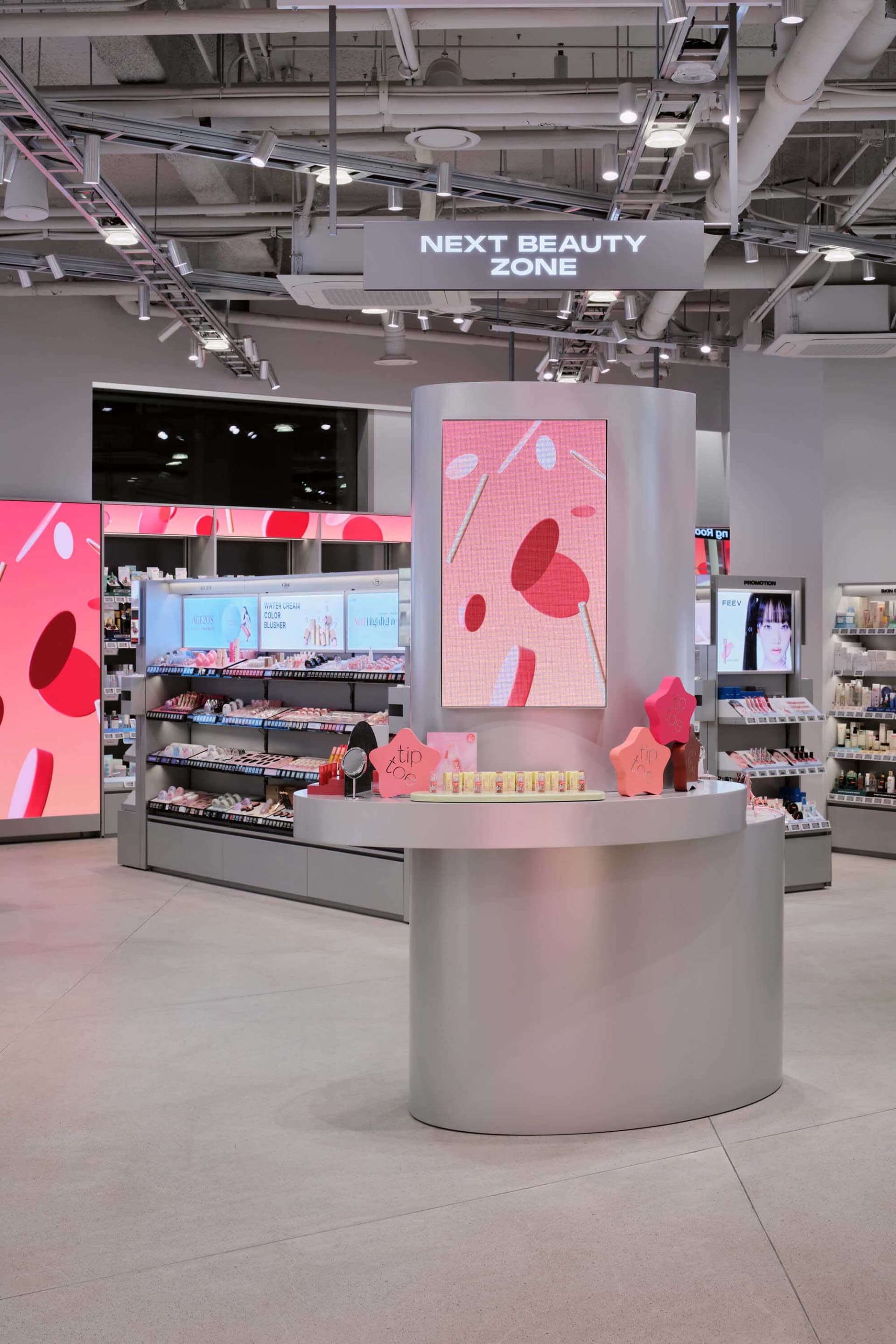 Modern silver curved beauty counter kiosk with large pink digital screen and product displays in a brightly lit retail space