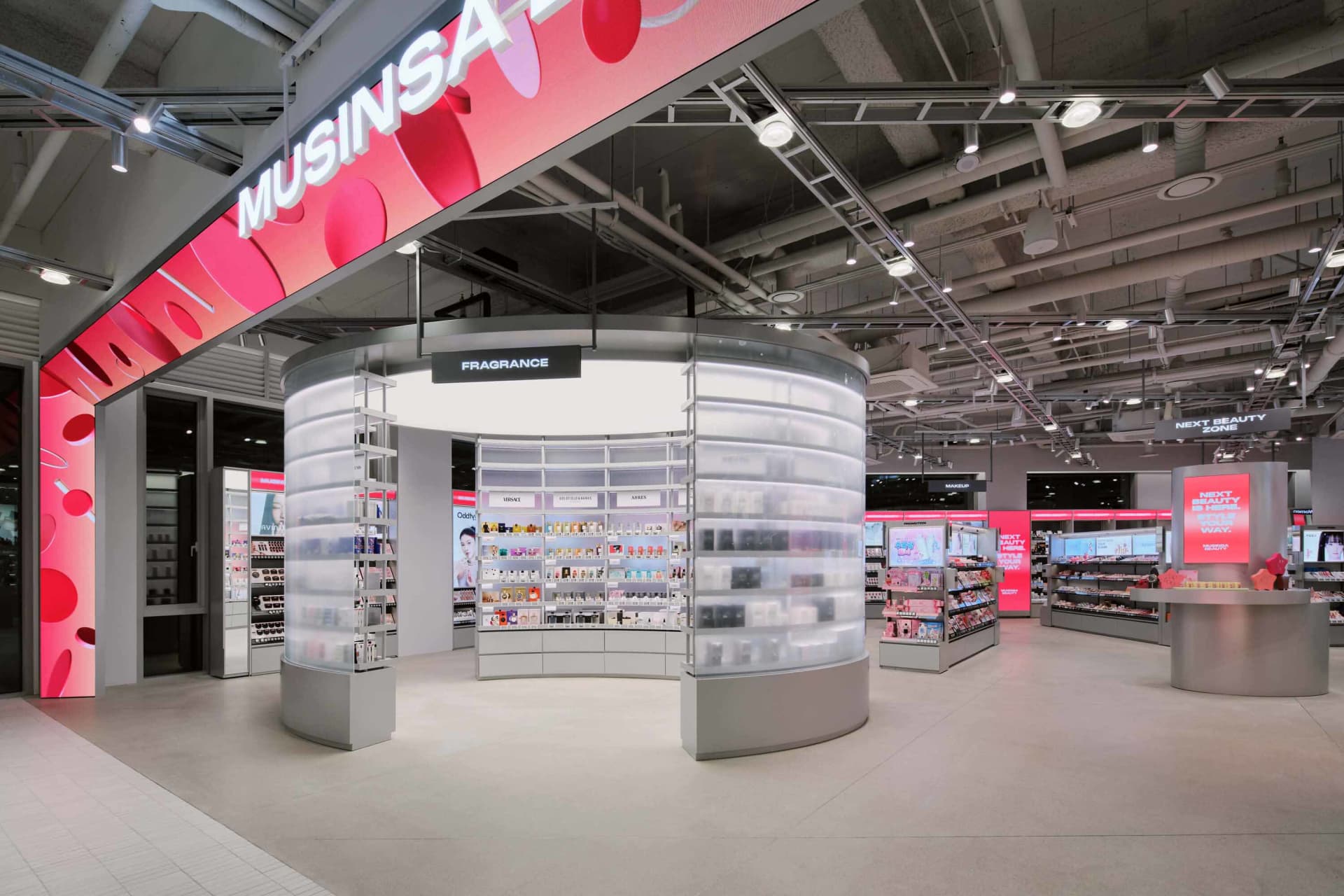 Modern beauty store interior with a large circular fragrance kiosk made of translucent acrylic shelves and integrated lighting, under a pink digital banner reading MUSINSARA.