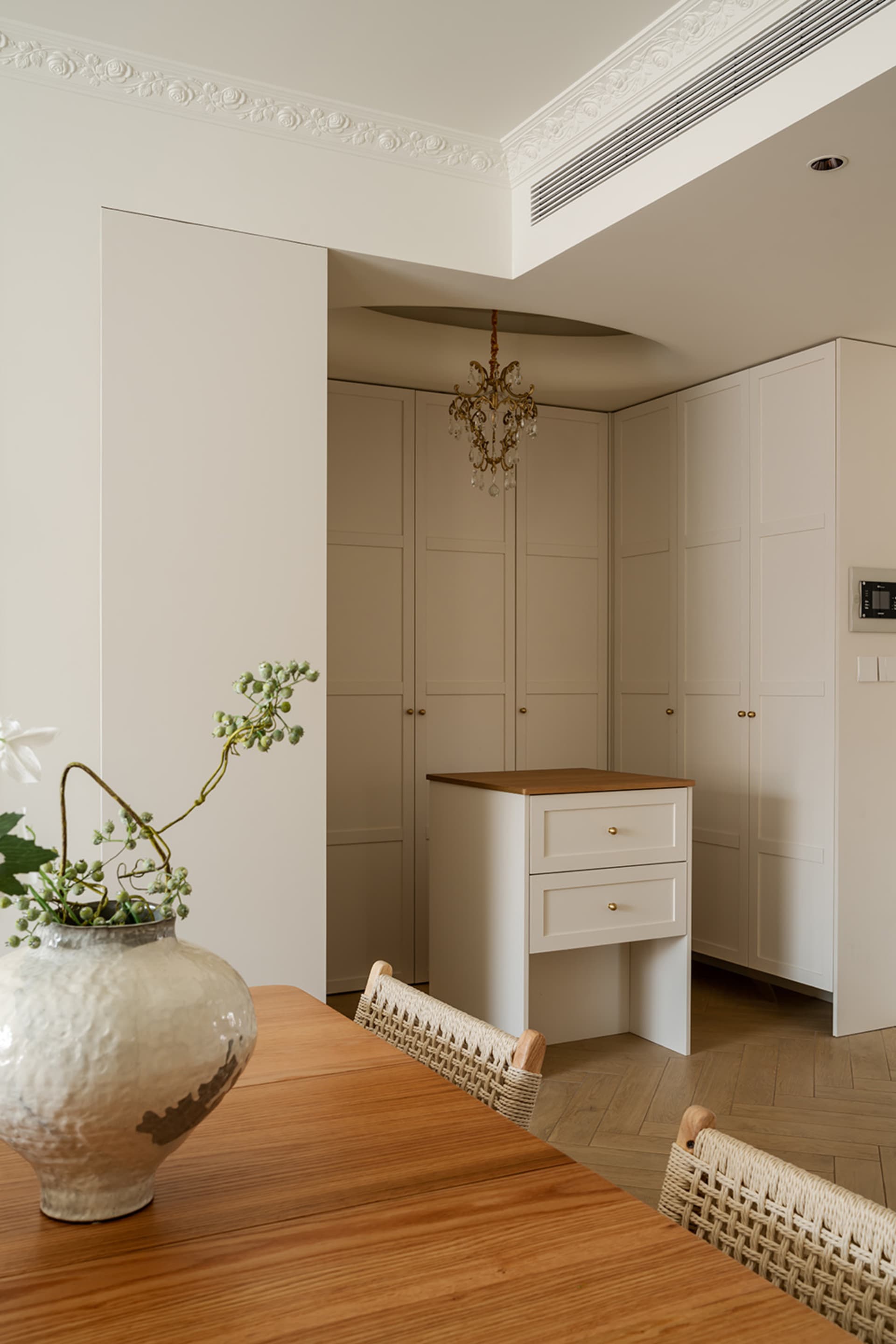 Elegant dining area featuring a wooden console table, woven rattan dining chairs, a large ceramic vase with greenery, and tall cream-colored built-in cabinetry with crown molding.