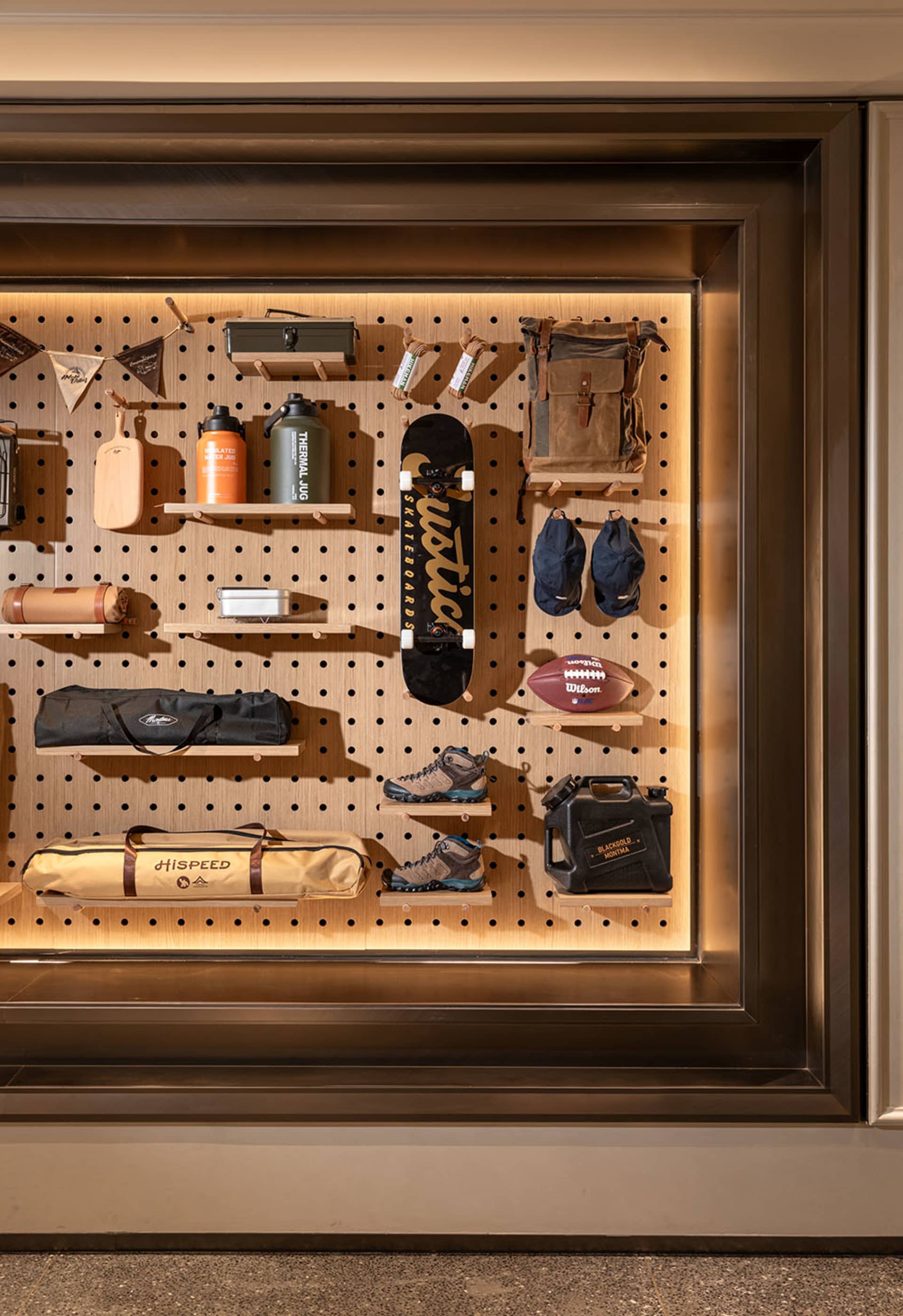 Modern recessed display wall featuring warm backlit oak pegboard with neatly arranged outdoor gear including skateboard, hiking boots, duffel bags, water jugs, and American football.