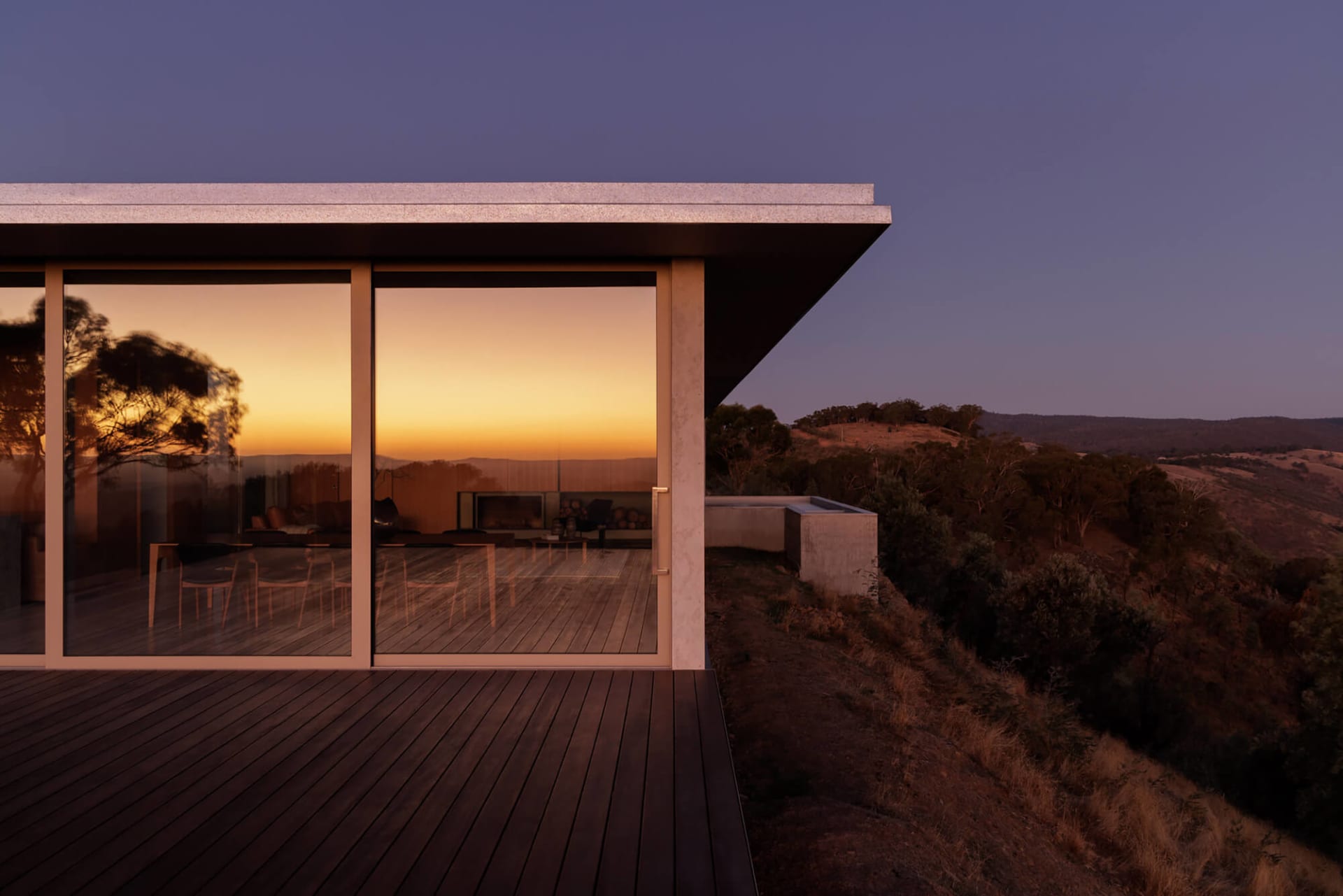 Modern hillside house with large glass windows and concrete overhang overlooking a golden sunset in a rural landscape