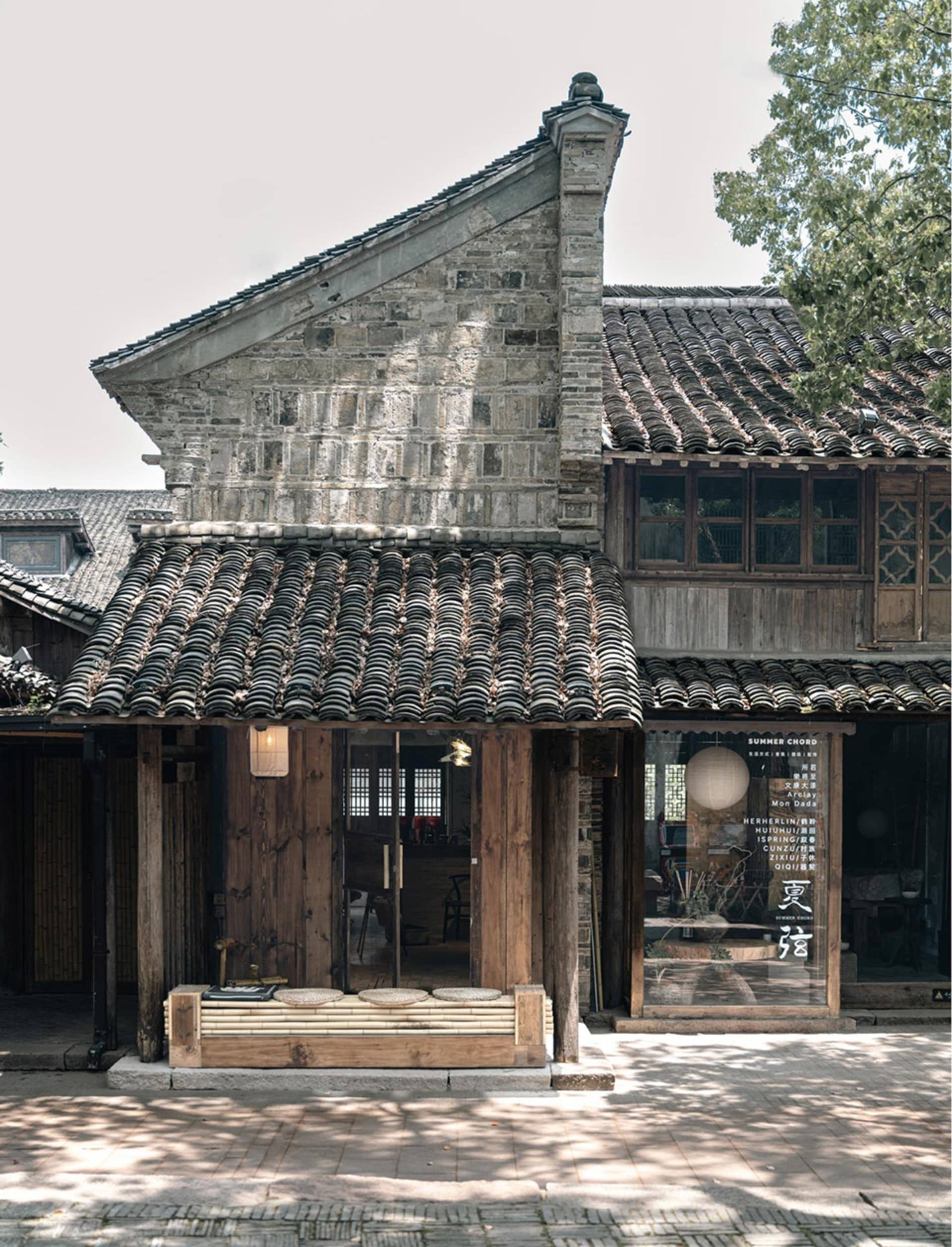 Exterior view of a traditional Chinese grey brick building with dark curved tile roofs, wooden pillars, and a bamboo bench at the entrance in Wuzhen.