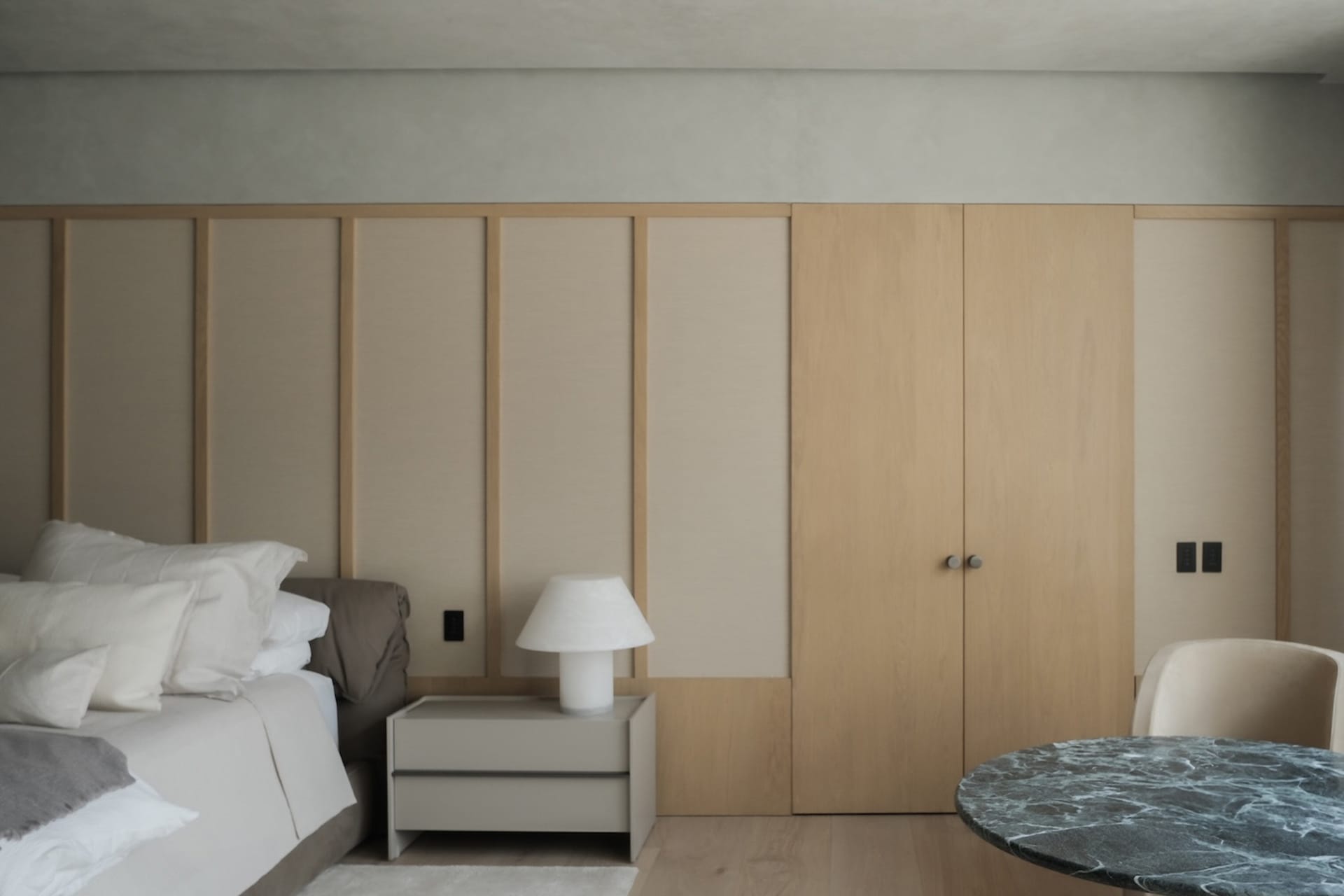Modern minimalist bedroom with light oak built-in wardrobe, beige upholstered headboard, white bedding, and a round green marble table in the foreground.