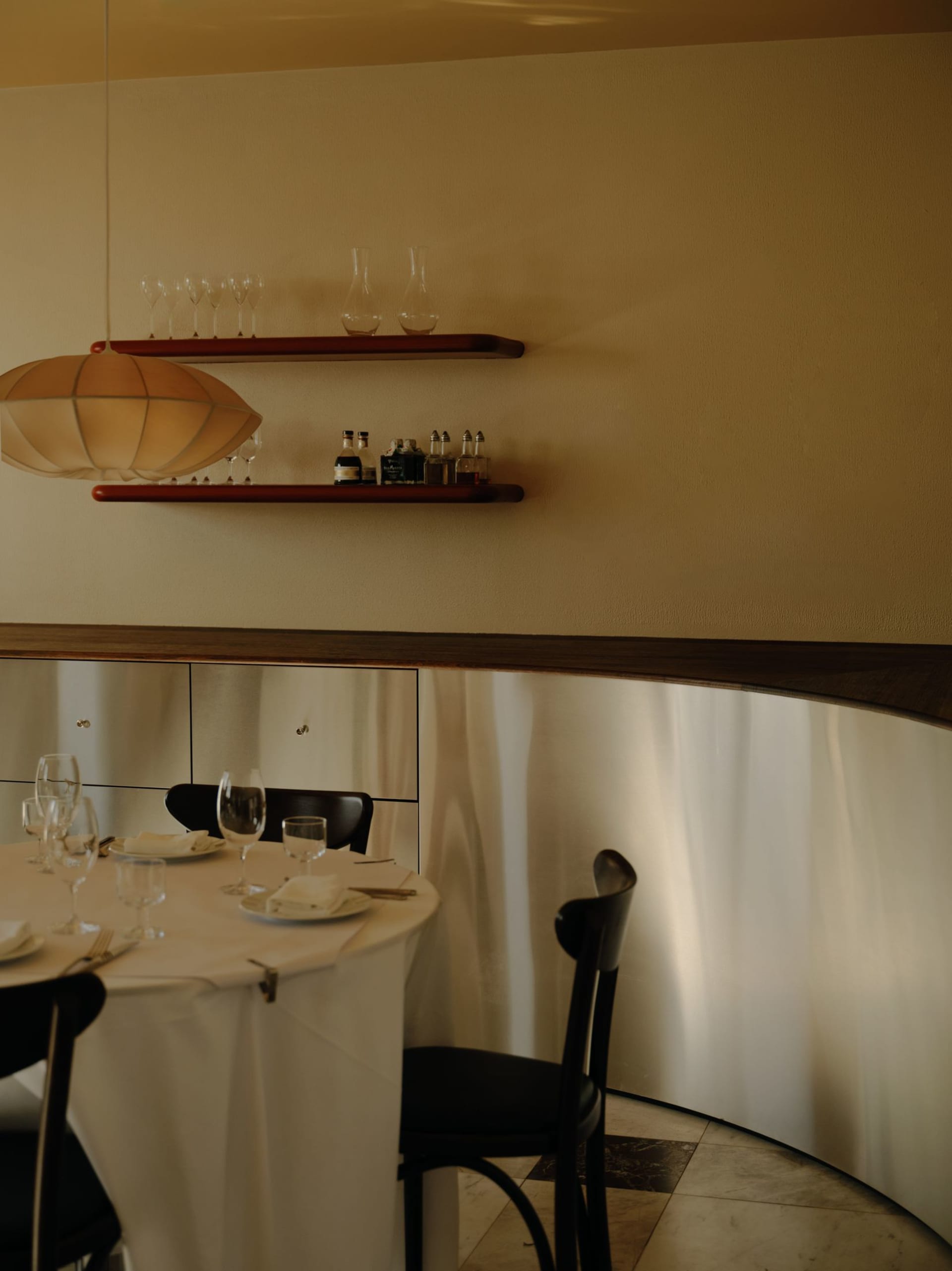 Elegant dining area featuring a curved walnut wall, built-in floating shelves, black dining chairs, and a round table set with white linens under a large pendant lamp.