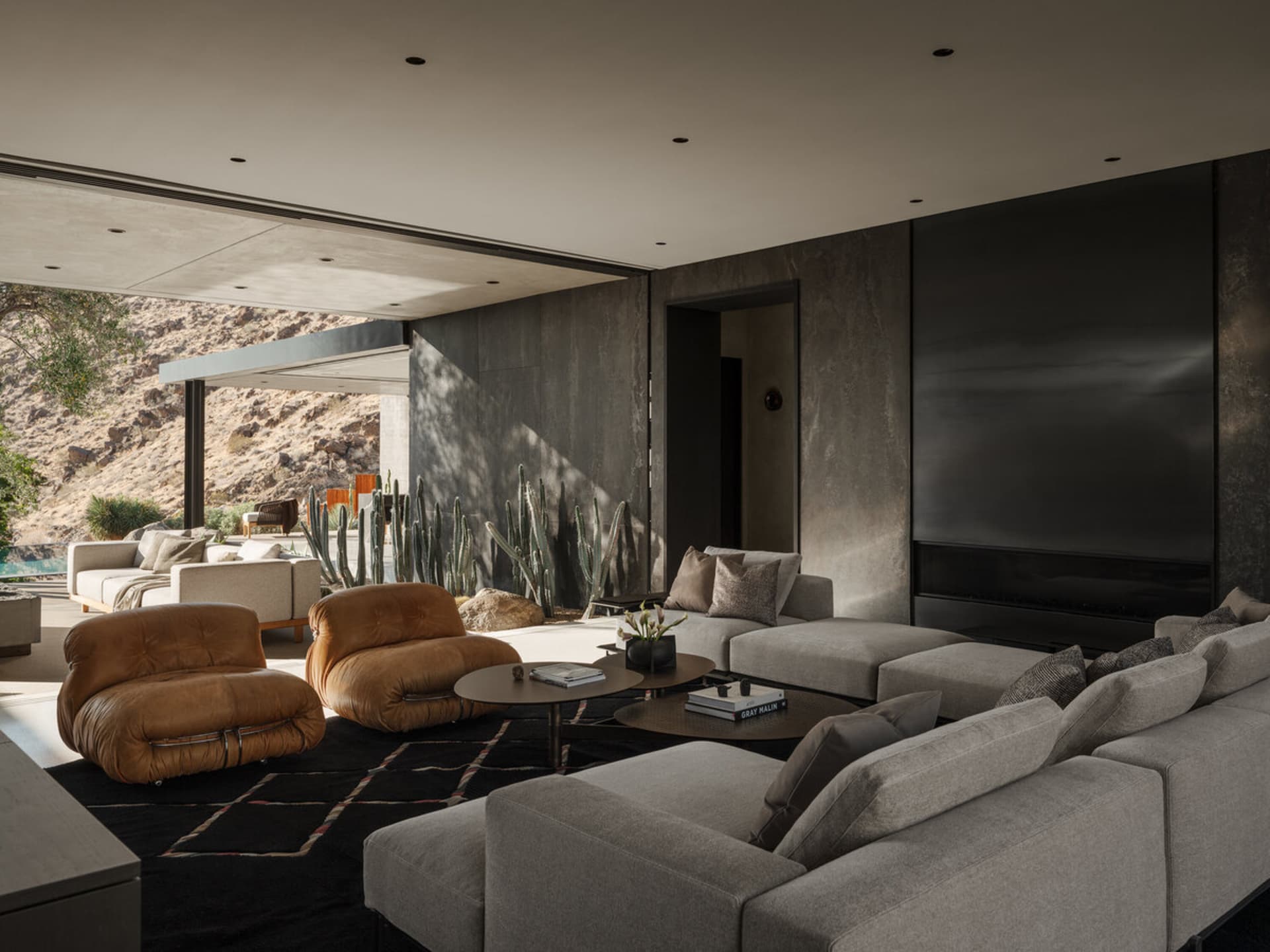 Modern desert living room featuring concrete walls, large grey sectional sofa, two cognac leather lounge chairs, round coffee tables on a dark rug, with desert landscape and cacti visible through open sliding doors.