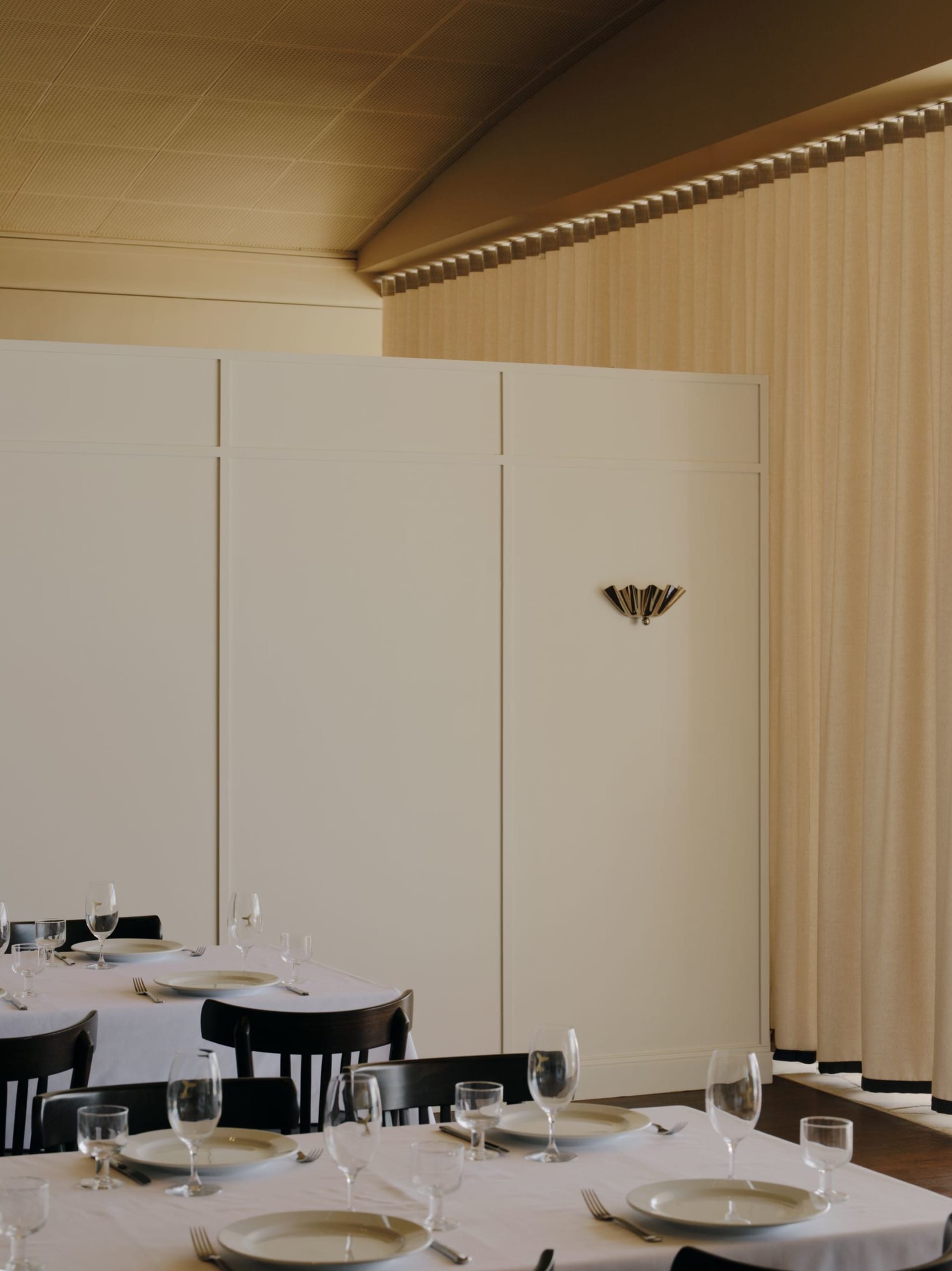 Elegant restaurant dining room featuring dark walnut chairs, white tablecloths, and tall cream curtains with a decorative brass wall sconce.