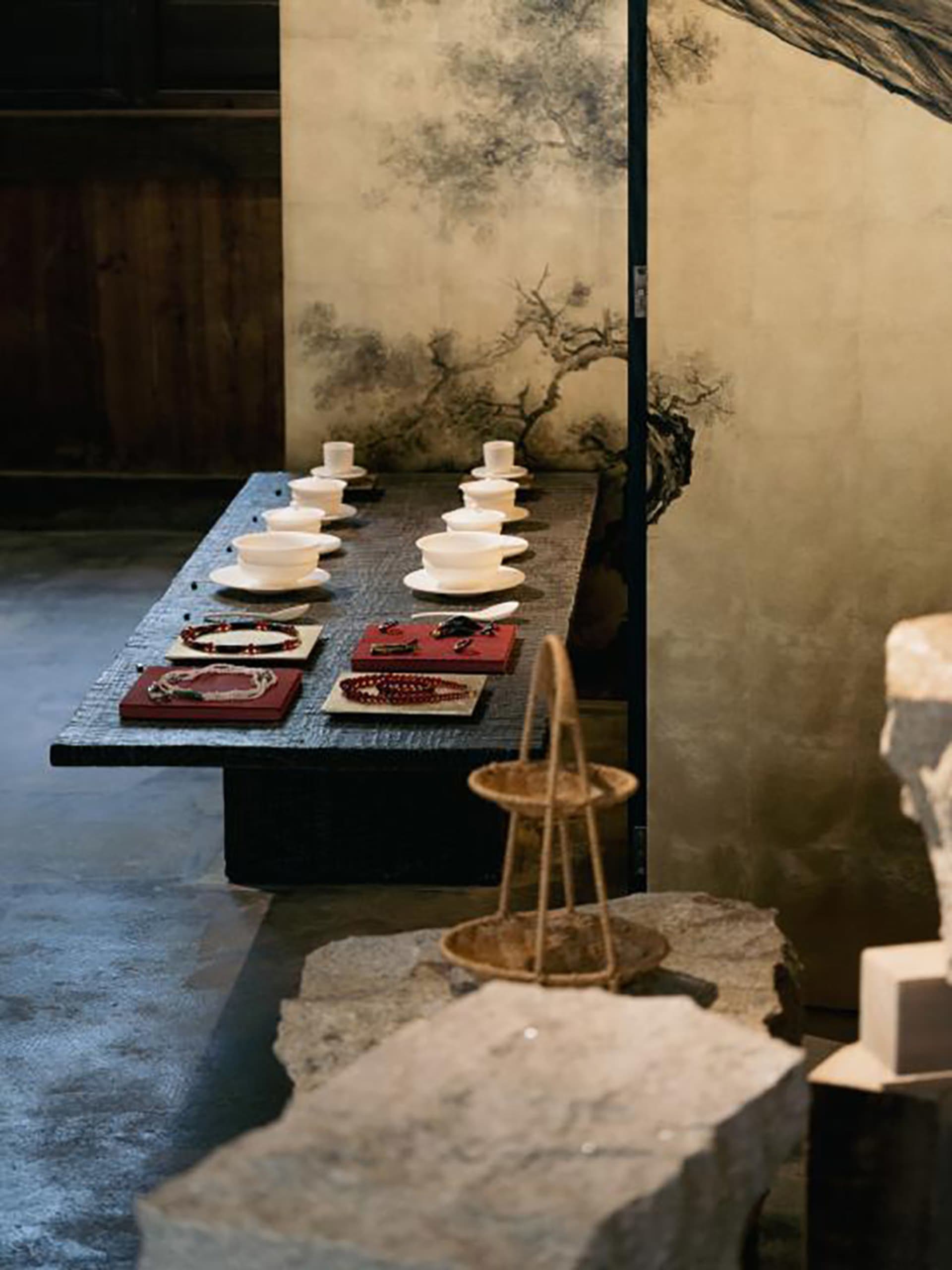Long dark stone table displaying white porcelain teacups and colorful beaded jewelry on red trays, set against a large Chinese ink-wash screen with a gnarled tree painting, in a minimalist wabi-sabi interior.