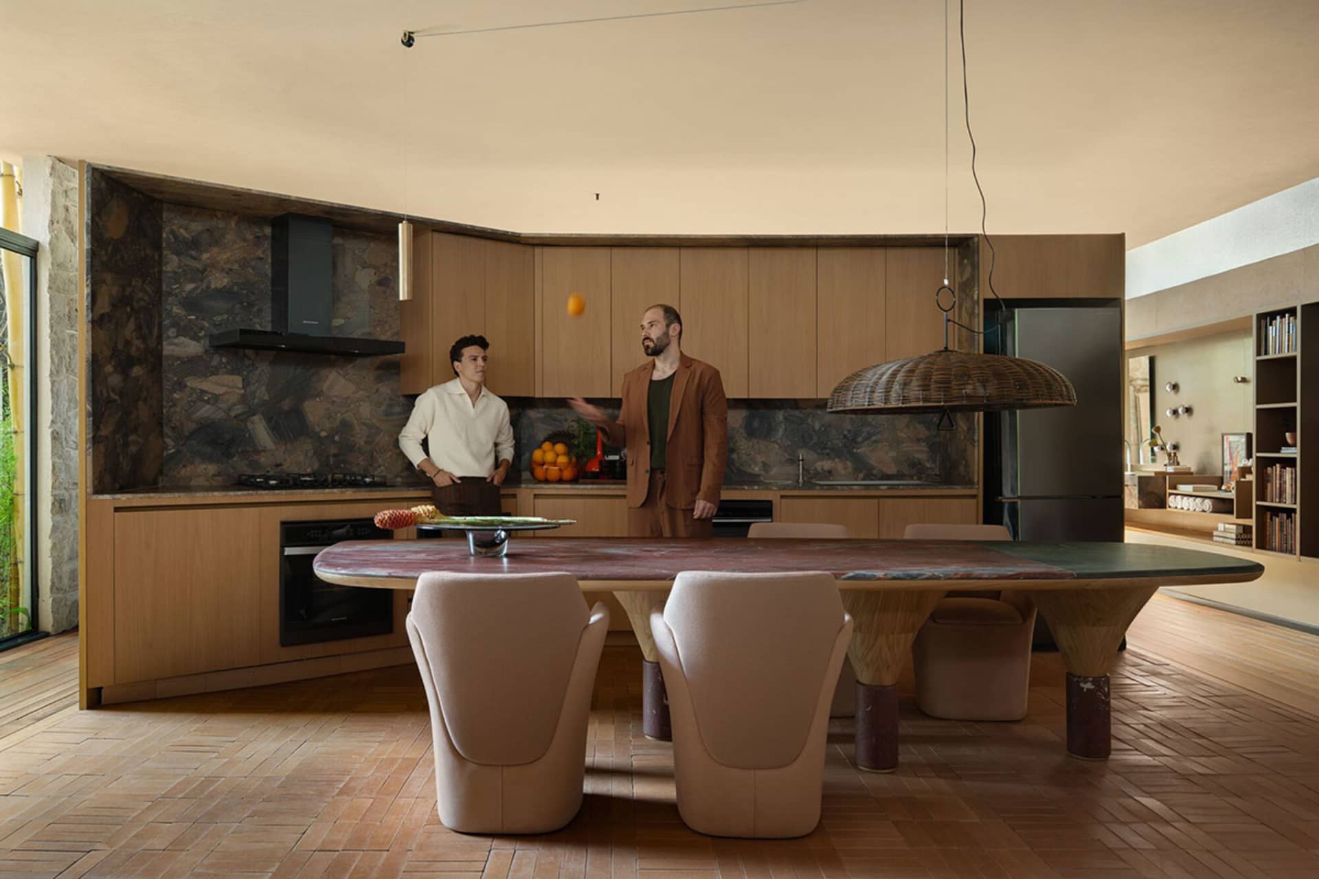 Modern open-plan kitchen and dining area with a large curved travertine dining table, sculptural wooden legs, beige dining chairs, light wood cabinetry, and a dramatic stone feature wall.