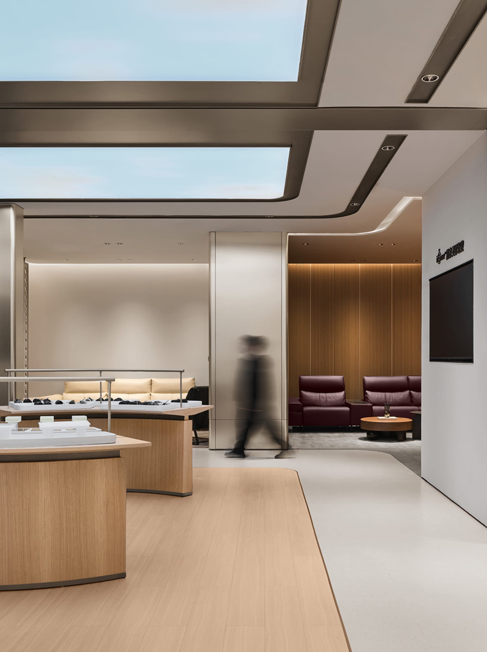Modern luxury jewelry boutique interior with curved oak display counters, beige sofas, burgundy leather armchairs, wooden slat accent wall, and large skylight.