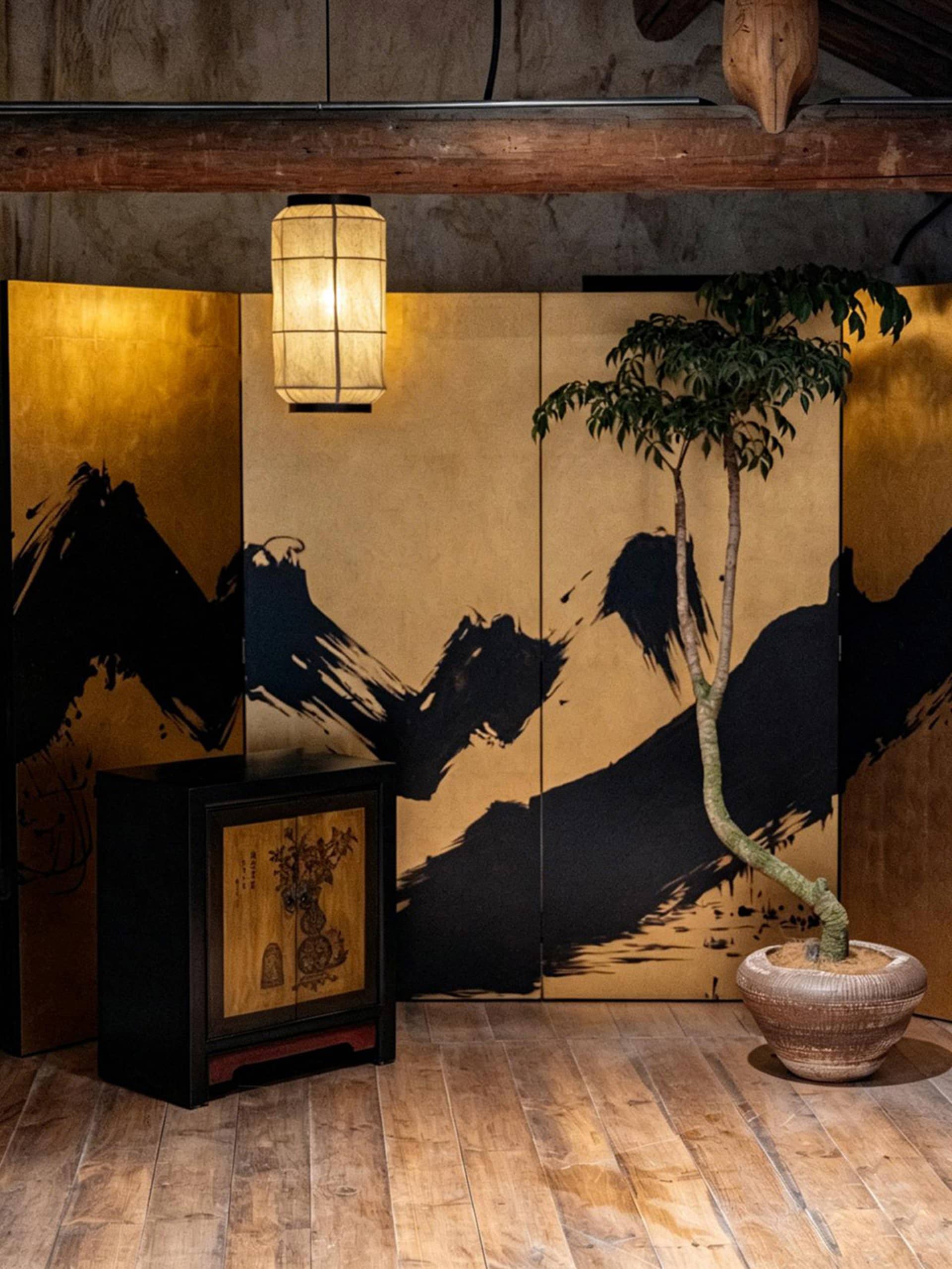 Japanese-style folding screen with black ink mountain landscape on gold leaf panels, accompanied by a black wooden cabinet, hanging paper lantern, and a tall bonsai tree in a ceramic pot on wooden flooring.