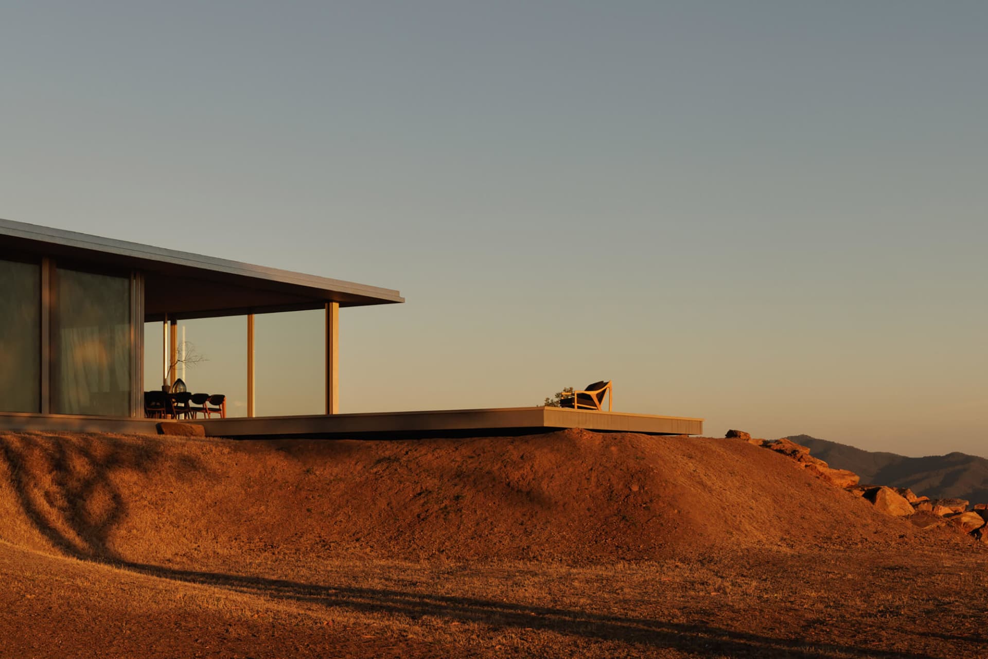 Modern minimalist house with large glass windows and concrete terrace overlooking a golden desert hill at sunset, with mountains in the distance.