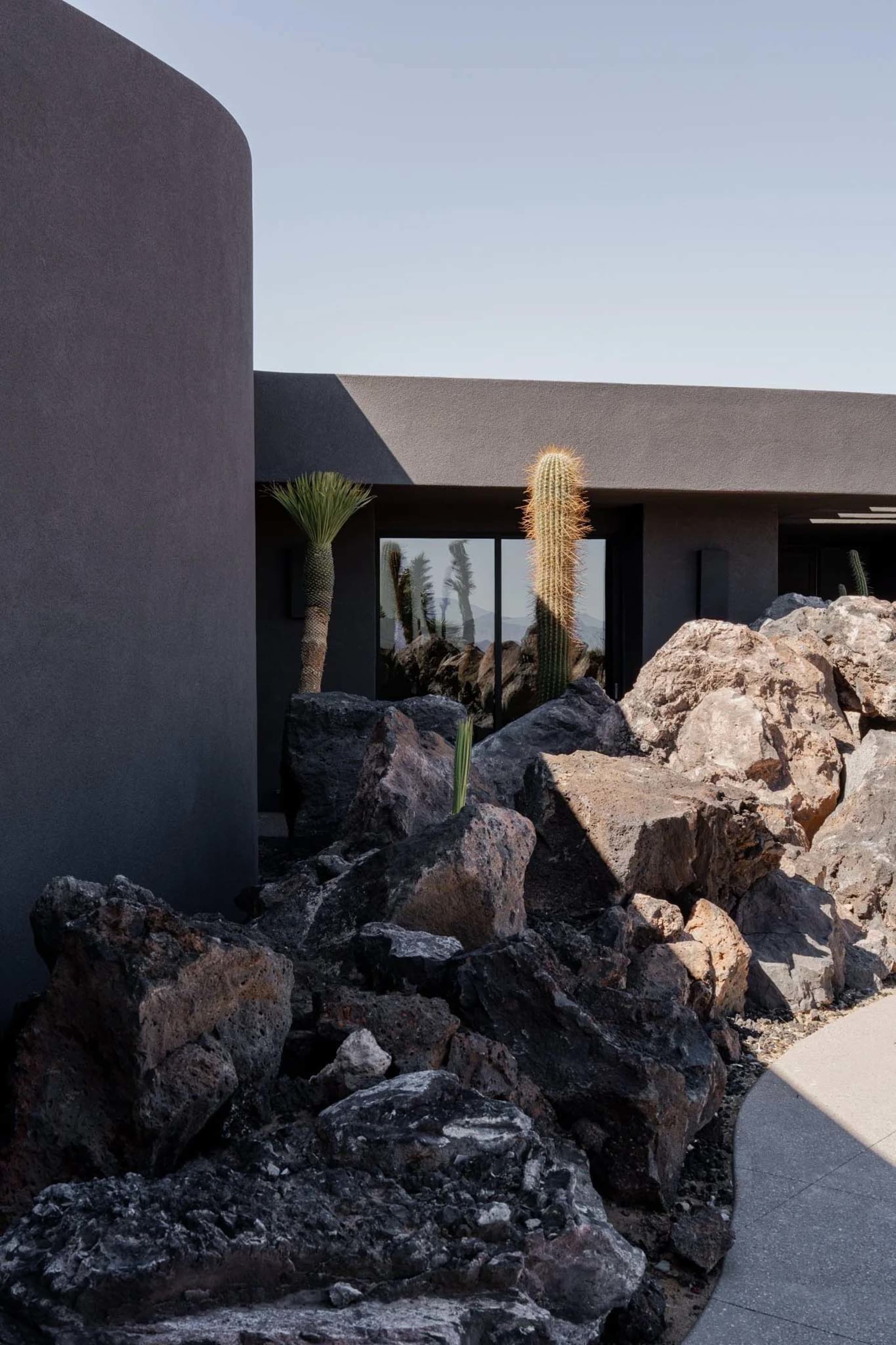 Exterior view of a modern desert house featuring dark curved concrete walls, large glass windows, and a foreground of dark volcanic rocks with saguaro and palm trees under a clear sky.
