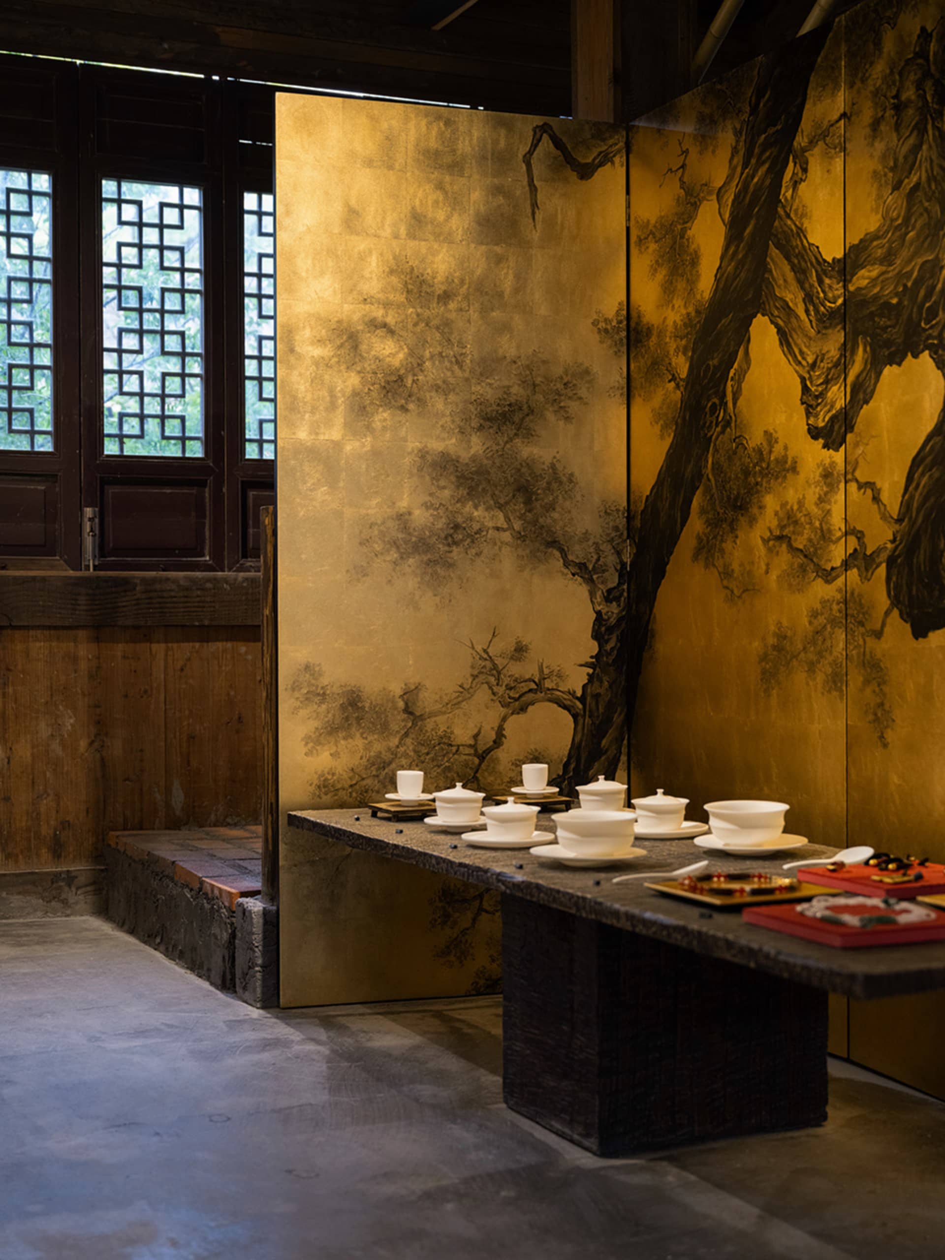 Interior view of a traditional Chinese tea room featuring a large gold-leaf folding screen with black ink pine tree paintings, a dark stone tea table with white porcelain tea ware, wooden lattice windows, and concrete floor.