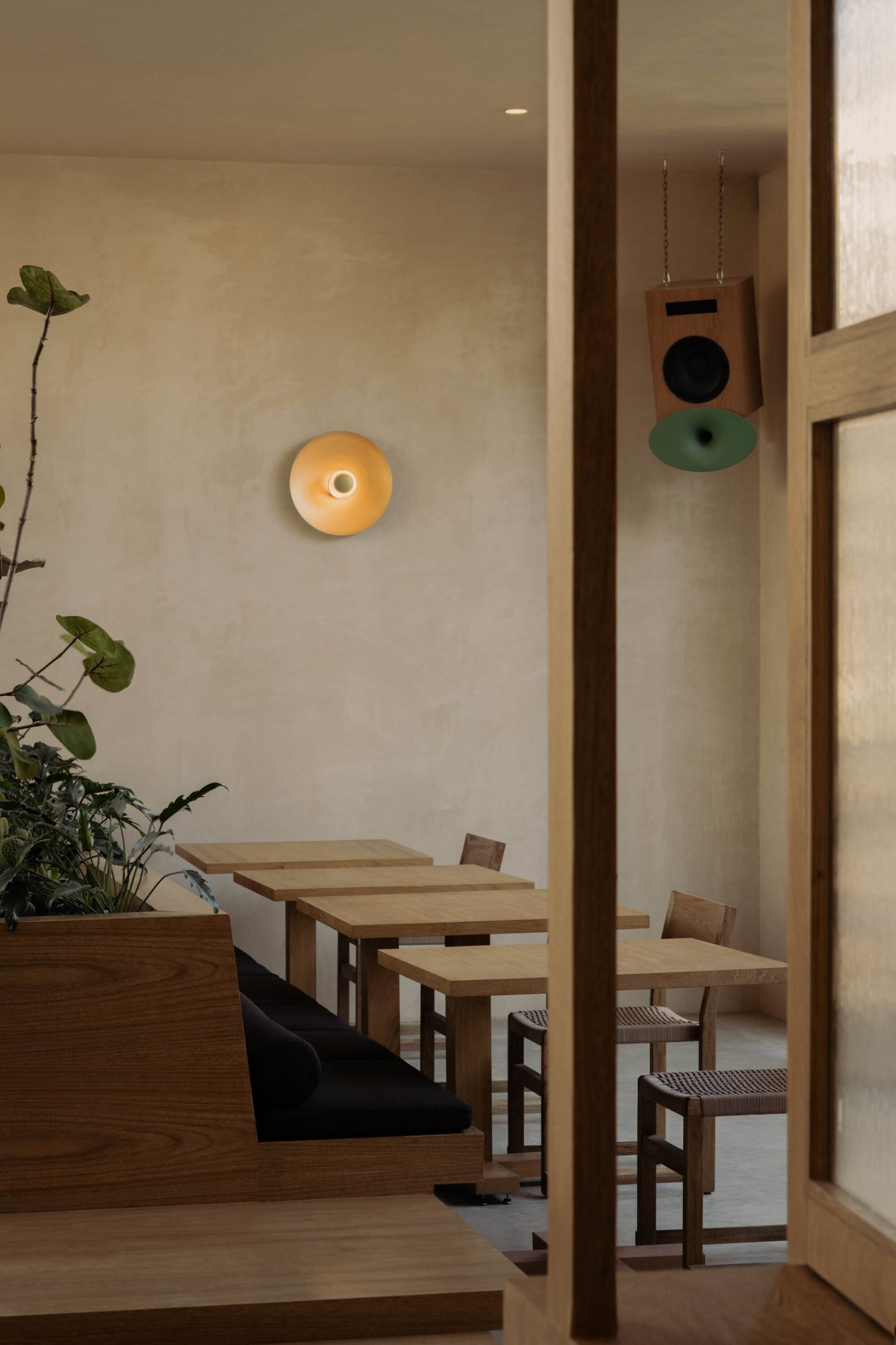 Minimalist cafe dining area featuring solid oak tables and chairs, a built-in wooden banquette with black cushions, indoor plants, a modern wall sconce, and a hanging wooden speaker with green cone.