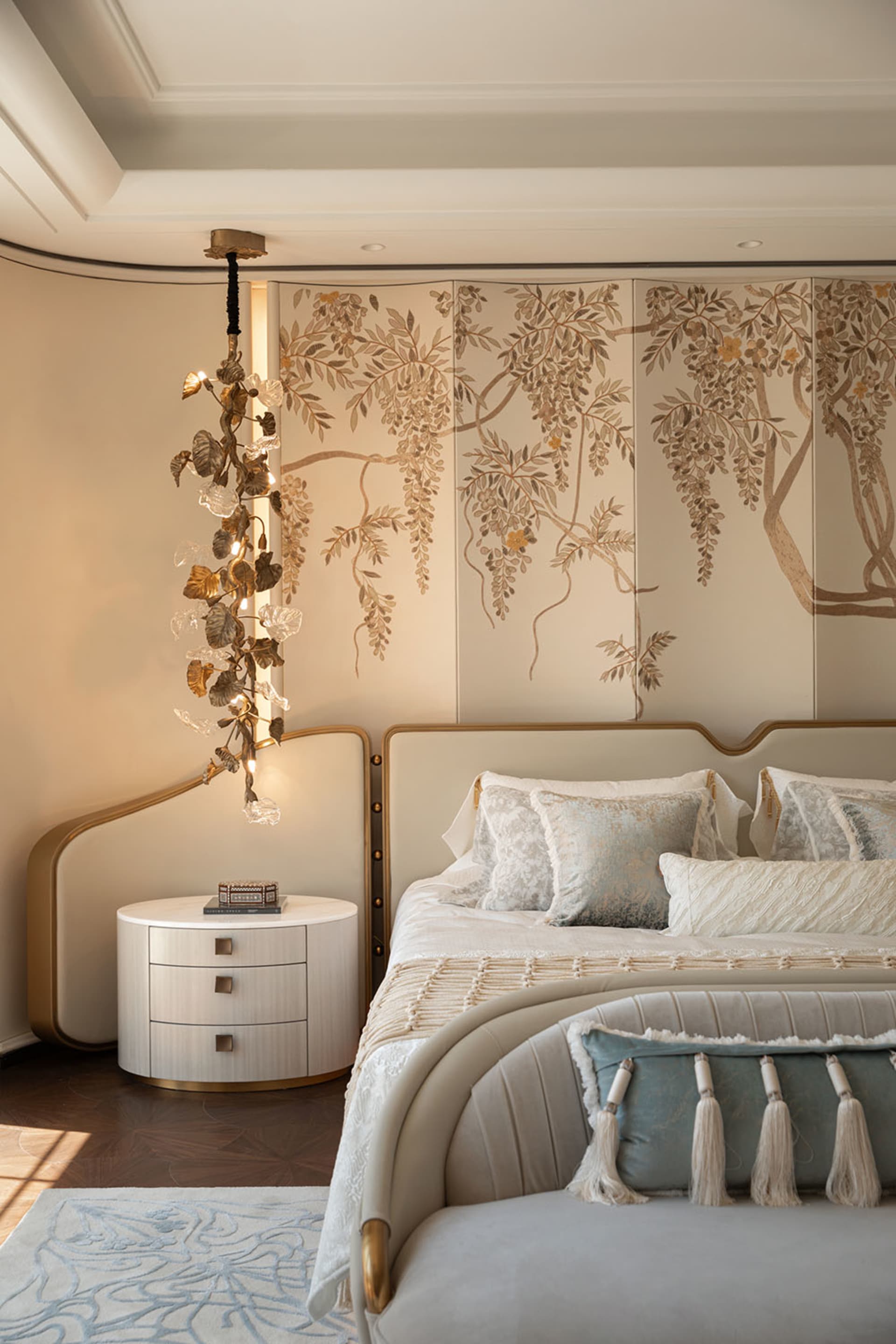 Elegant modern luxury bedroom with a large Chinese-style folding screen painted with wisteria flowers, a sculptural brass and crystal chandelier, curved upholstered bed with blue and cream pillows, round white nightstand, and light blue patterned rug.