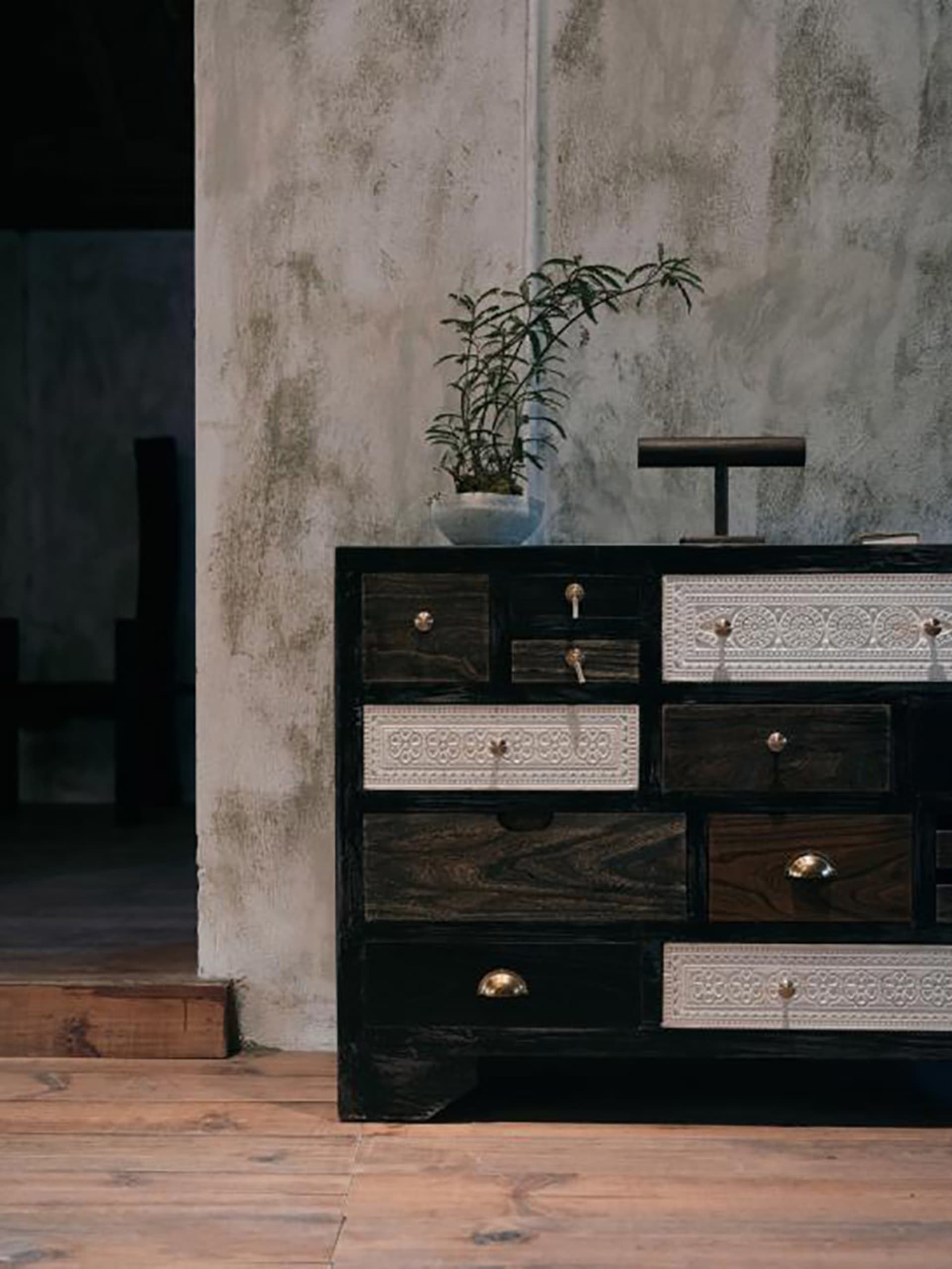 Vintage-style black reclaimed wood sideboard with white carved drawers and brass pulls, placed against a textured concrete wall with a potted plant on top.