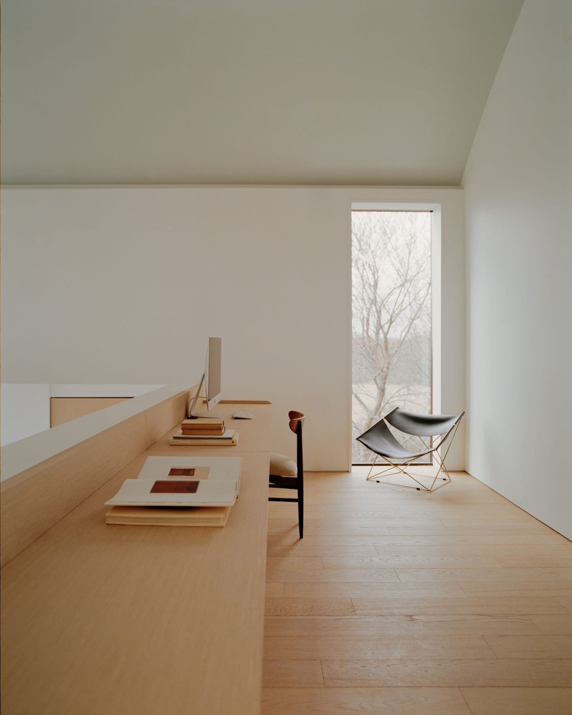 Minimalist home office featuring a long oak built-in desk, computer monitor, wooden chair, and two black butterfly chairs by a large window with natural light and bare trees outside.