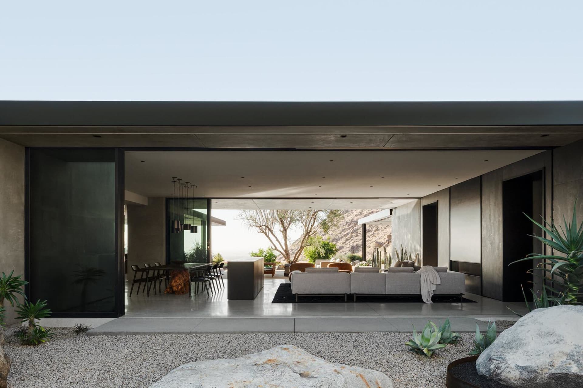 Modern desert house interior with large concrete volumes, open-plan living and dining area, and expansive mountain views through floor-to-ceiling glass.
