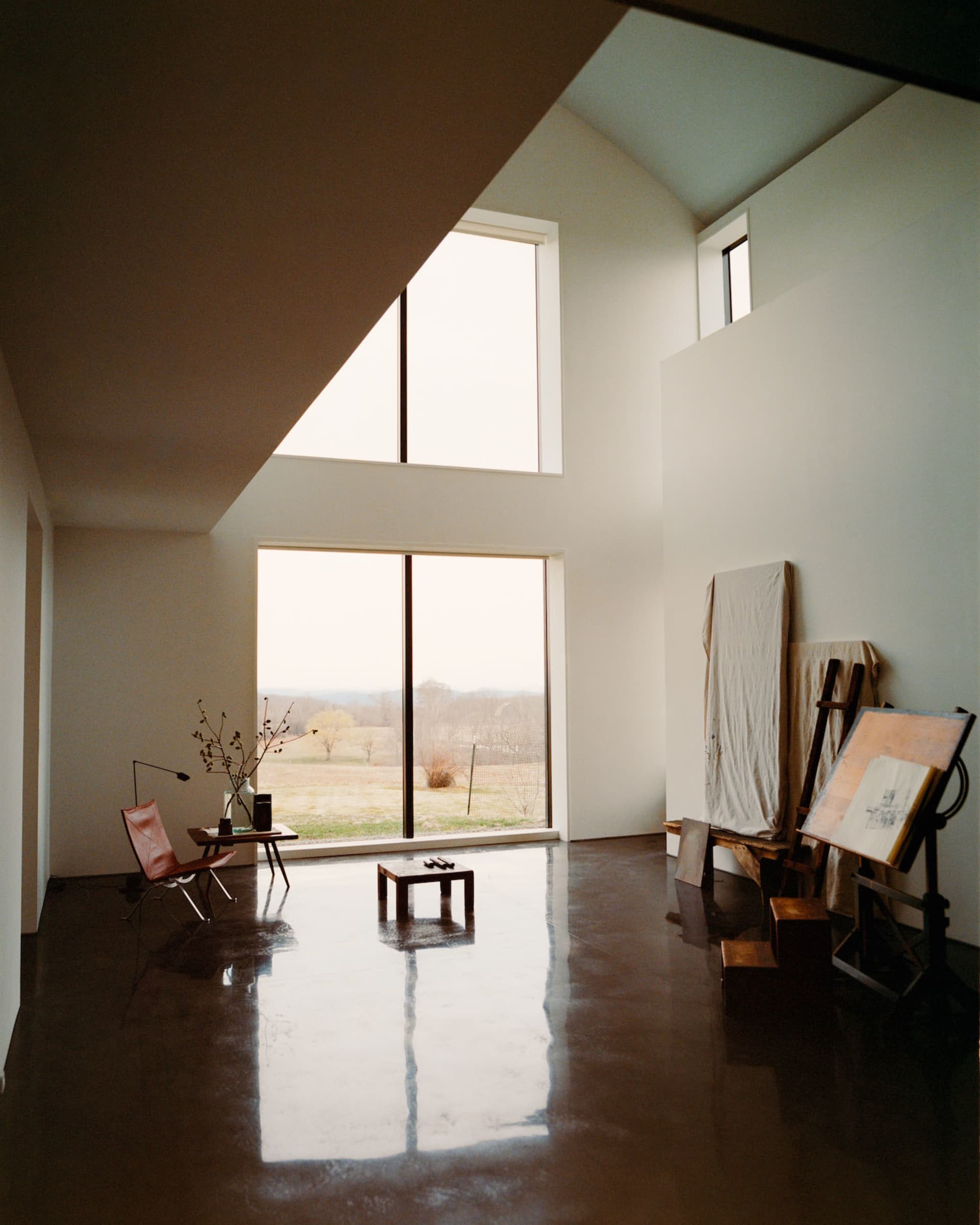 A spacious minimalist artist studio featuring a high cathedral ceiling, polished concrete floors, a leather butterfly chair, wooden side table with dried branches, and large easels with canvases on the right side.