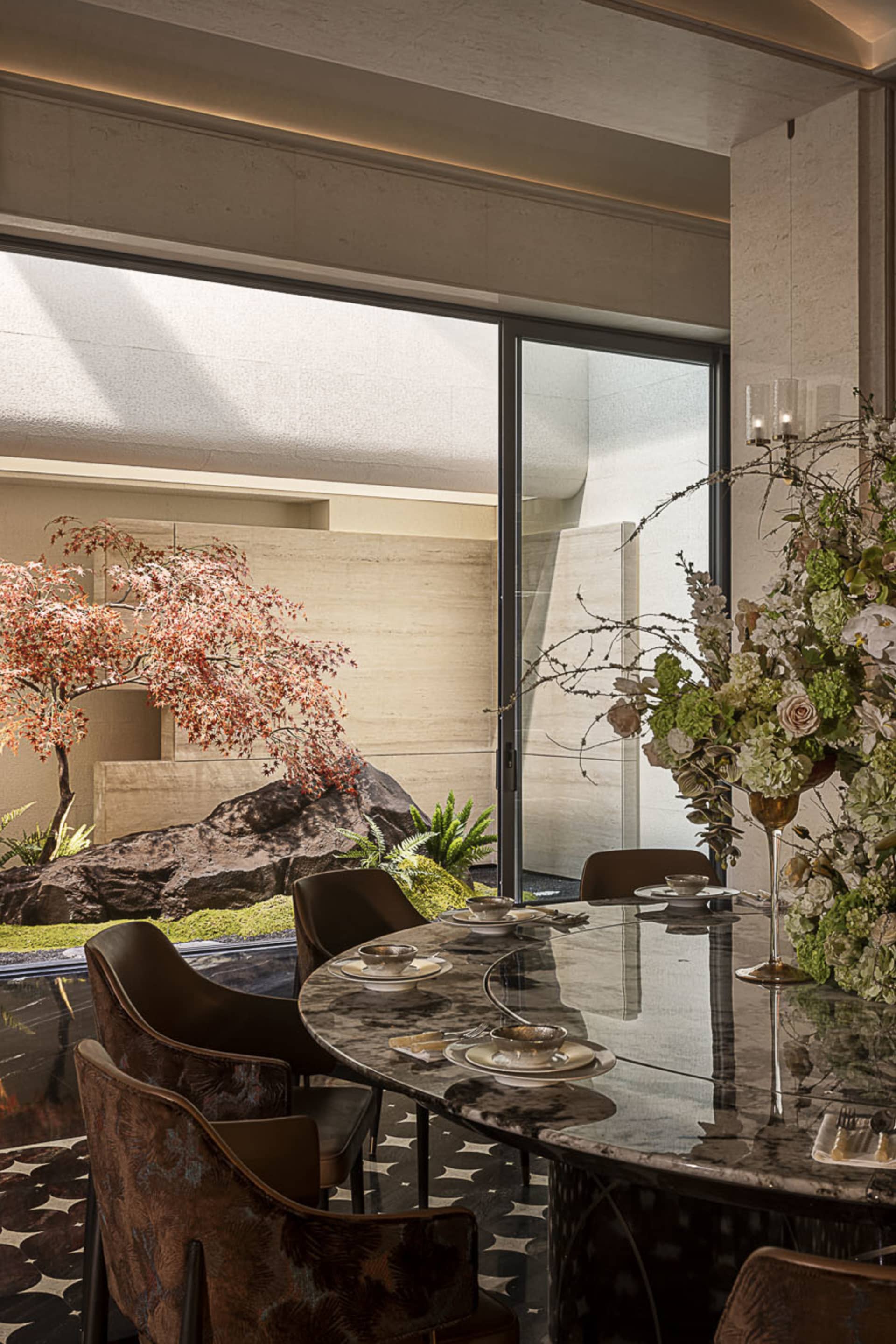 Modern luxury dining room featuring a dark marble oval table, velvet patterned dining chairs, and a view of a Japanese-style garden with red maple tree and large rocks.