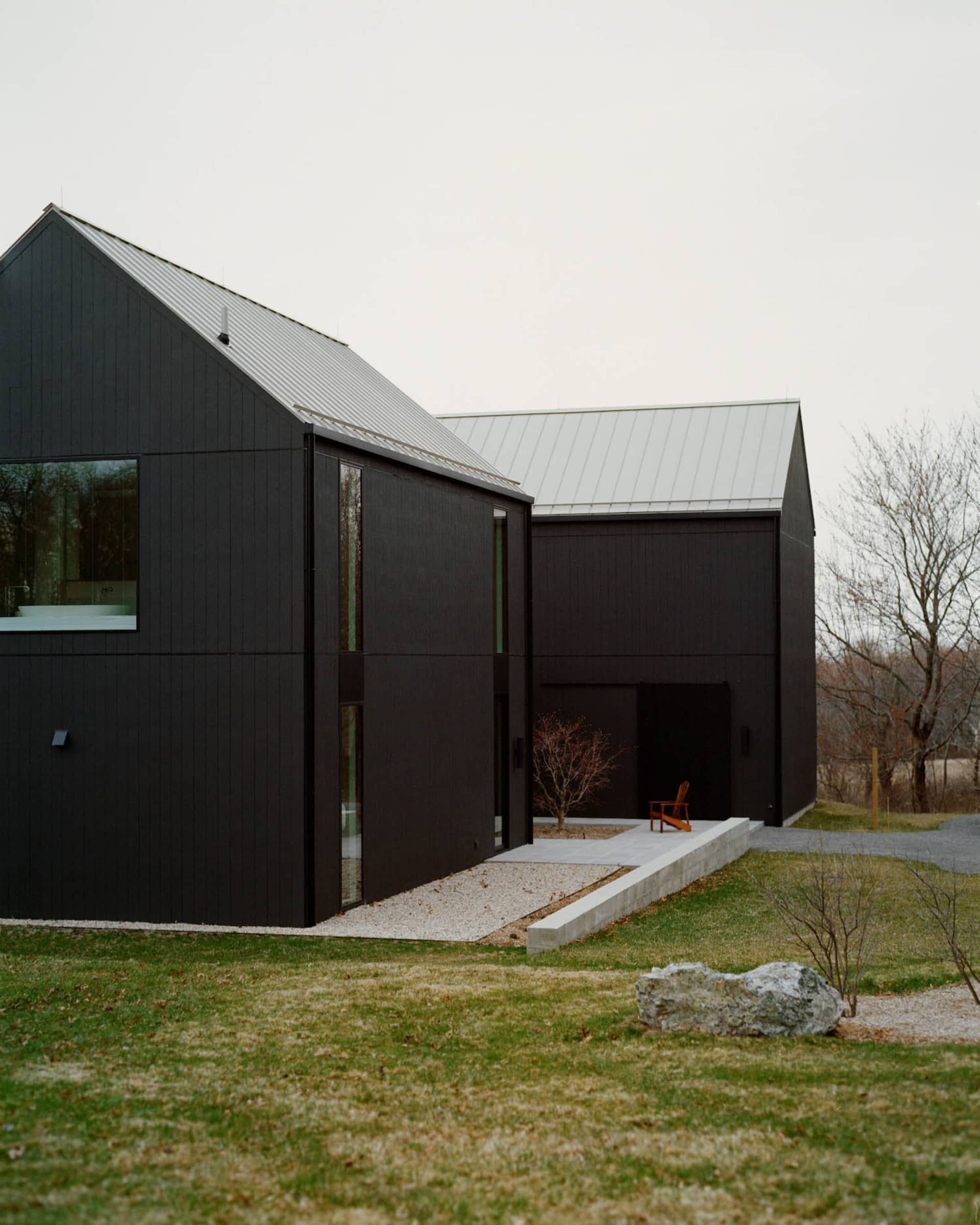 Modern black barn house with vertical wood cladding, large windows, metal roof, concrete terrace and gravel path set in a rural landscape.
