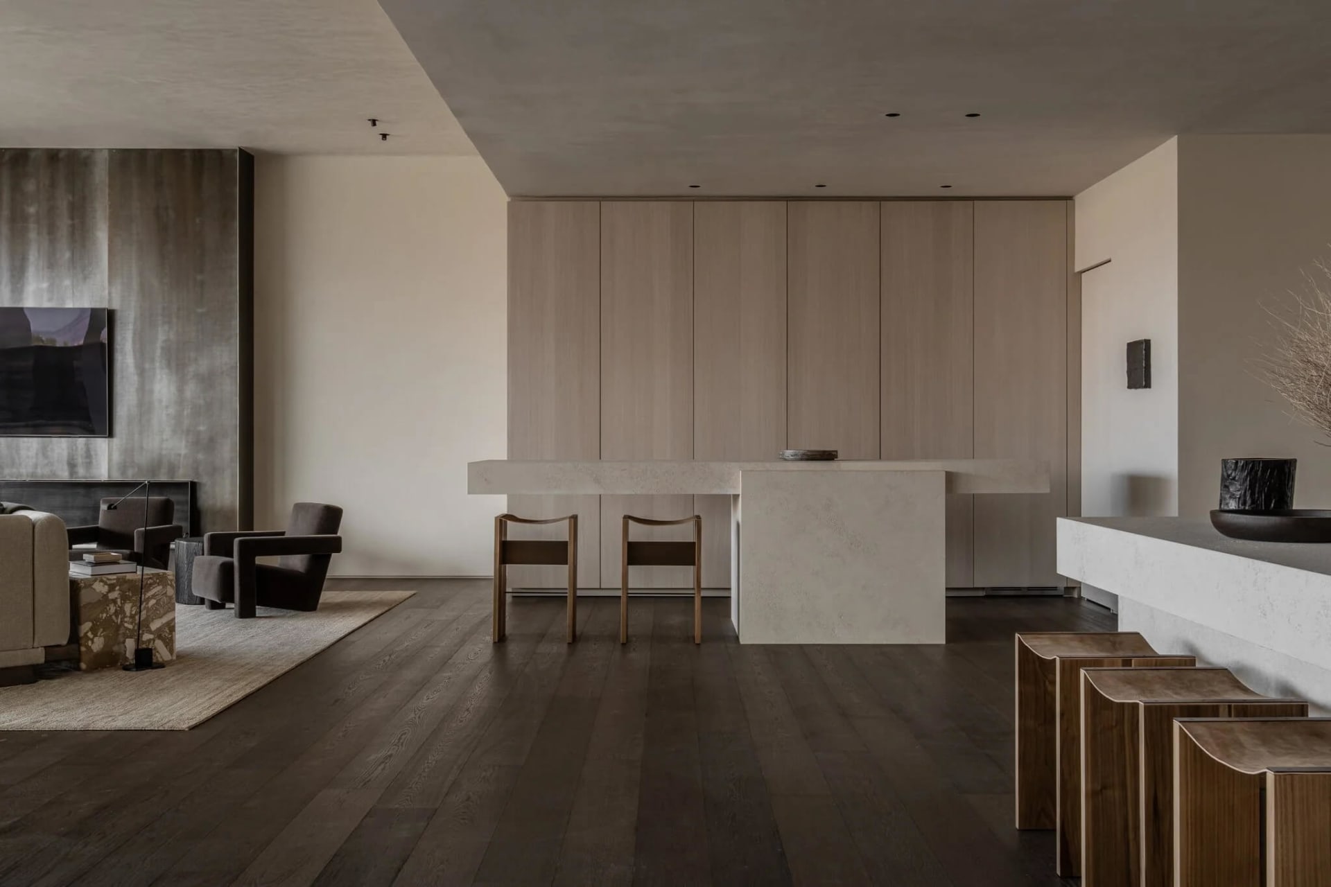 Modern minimalist open-plan living, dining and kitchen with concrete island, light oak cabinetry, dark oak flooring and sculptural wooden stools.