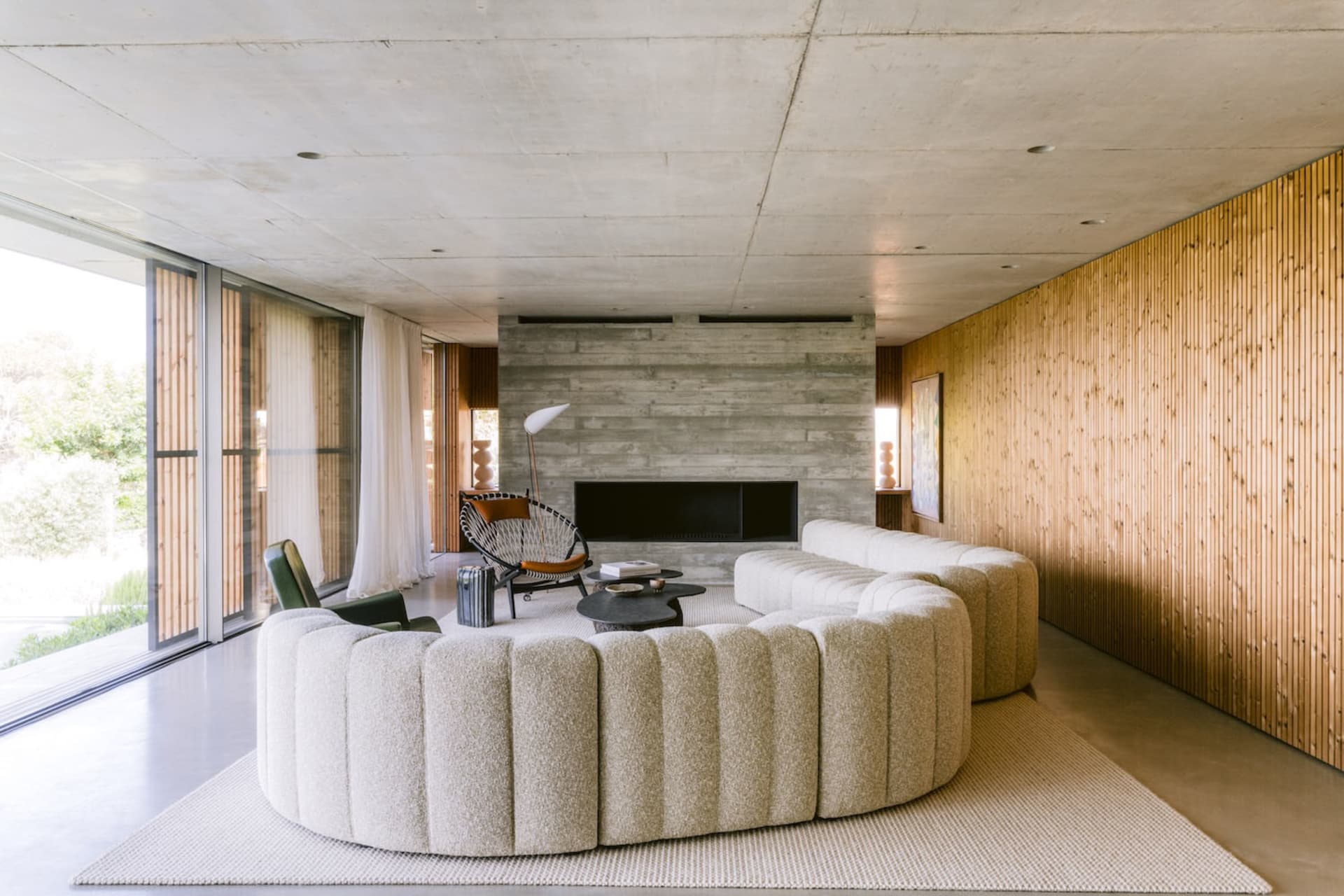 Modern living room featuring a large curved boucle sofa, concrete walls, vertical wood paneling, and a black woven accent chair in front of a fireplace.