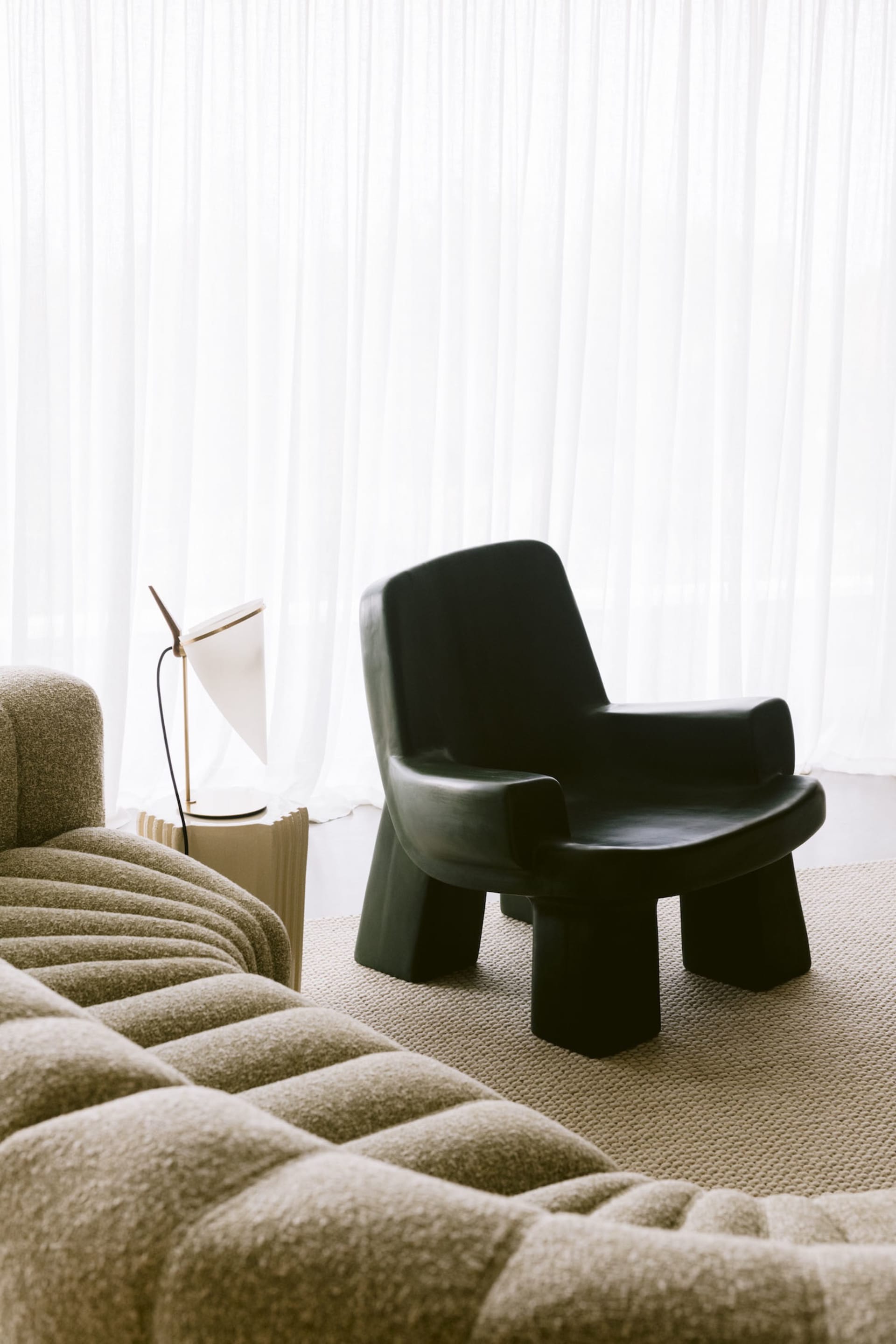 Modern living room corner featuring a black sculptural armchair, a beige boucle sofa, a white table lamp on a fluted side table, and floor-to-ceiling sheer white curtains.