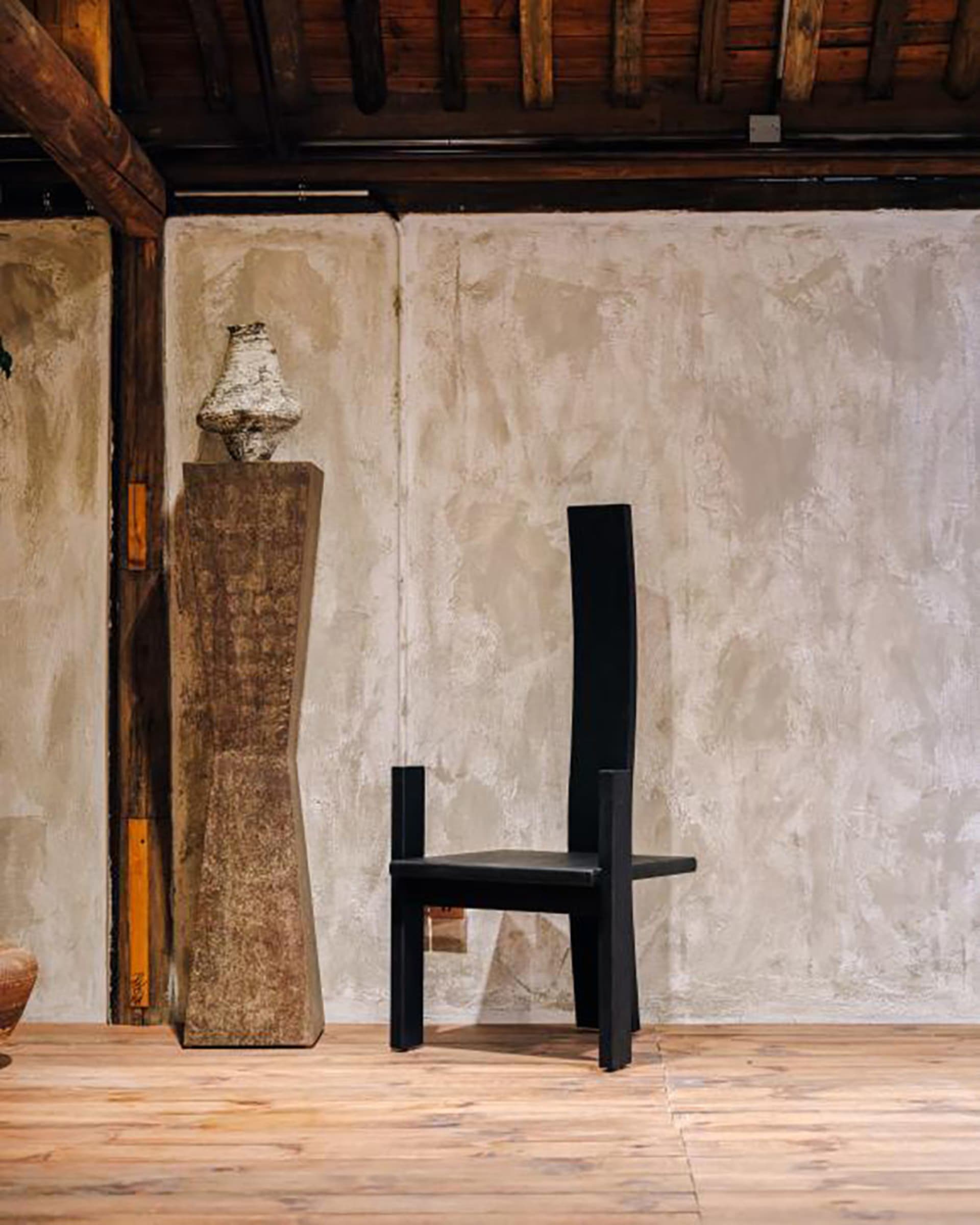 A tall black wooden chair with a straight back and angular frame placed on a light wood floor in front of a textured off-white plaster wall, accompanied by a stone pedestal with a ceramic sculpture.