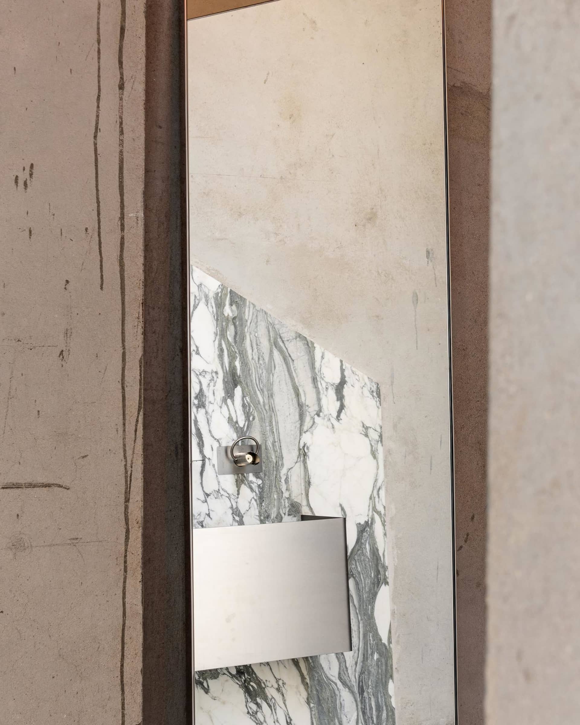 Close-up of a modern bathroom vanity mirror with white and green marble panel, stainless steel accessories, and raw concrete wall surround.
