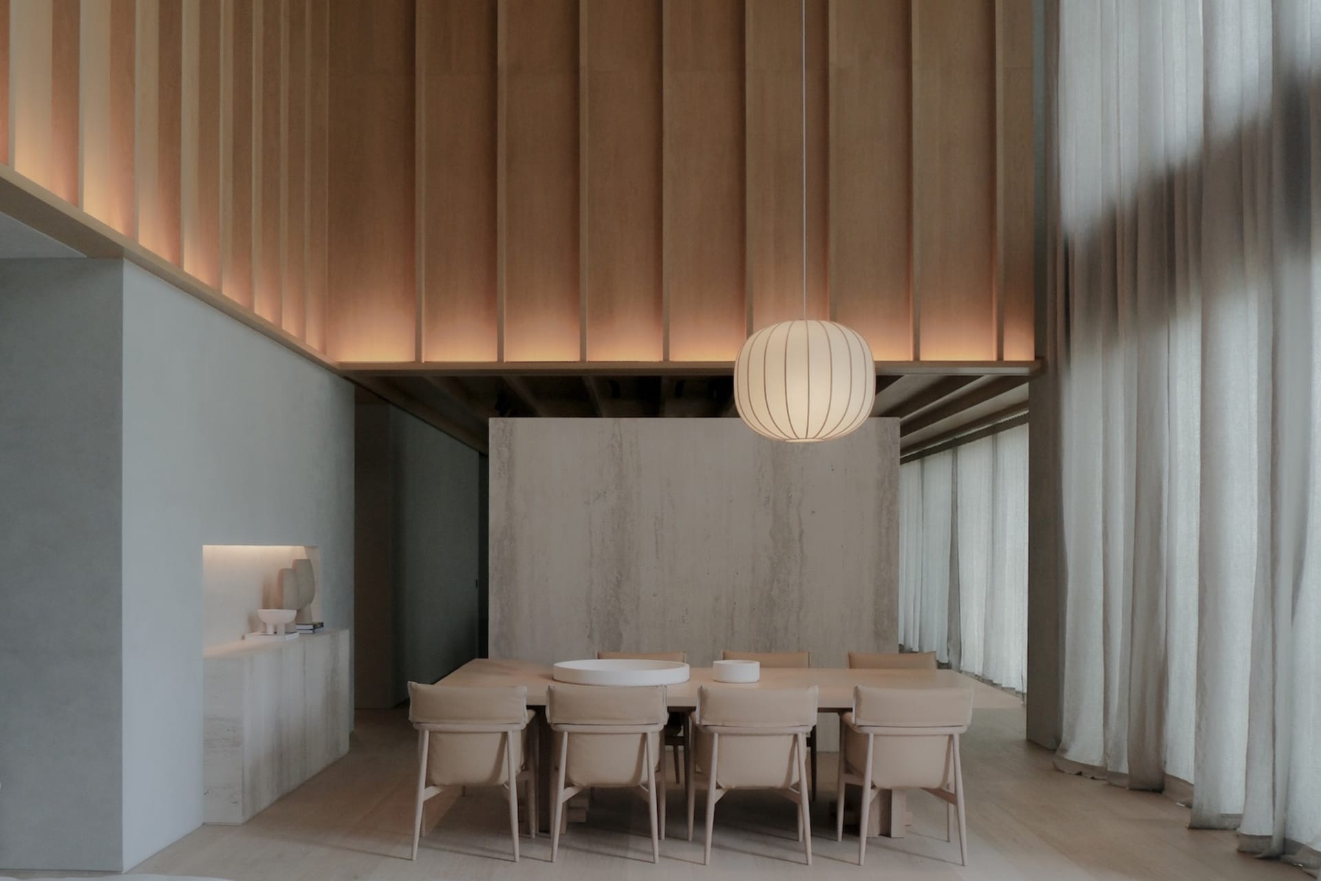 Modern minimalist dining room featuring a long oak dining table with beige upholstered chairs, a large ribbed pendant lamp, vertical wood slat ceiling, and a prominent concrete accent wall.