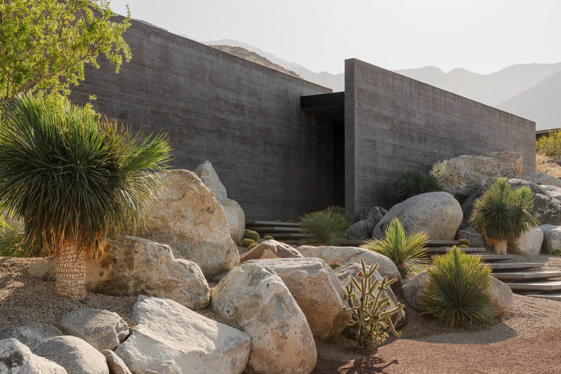 Modern concrete building with large boulders and desert plants including yuccas and cacti in an arid mountain landscape.